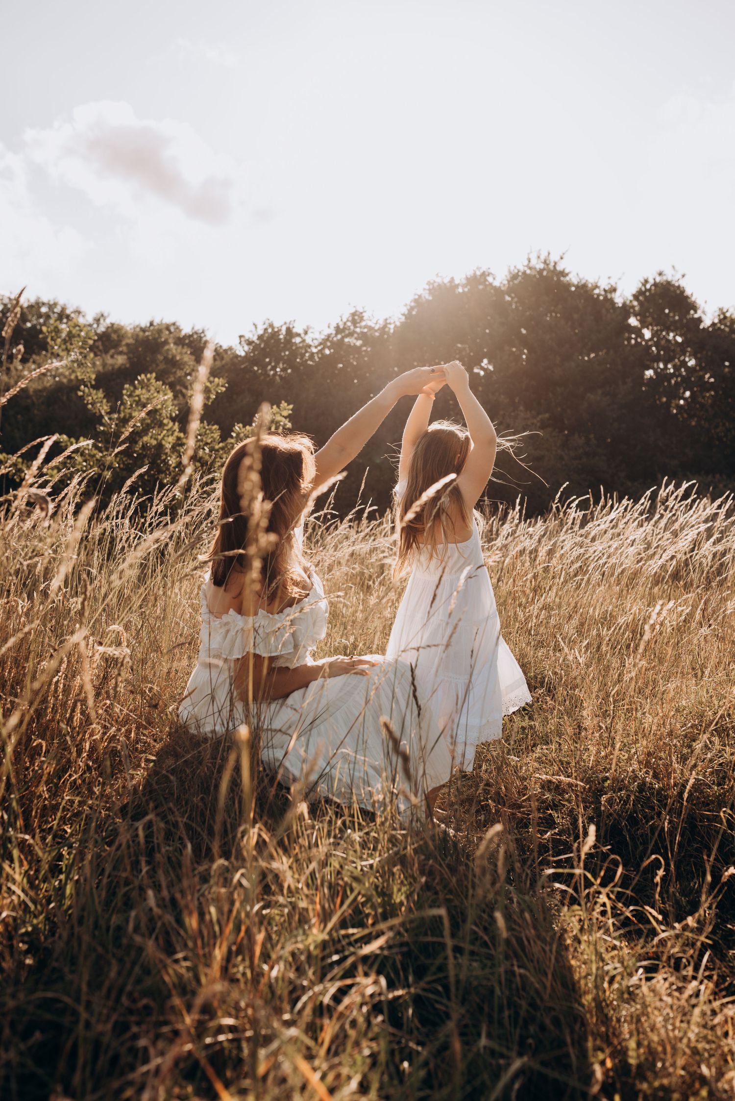 Two people in white dresses dance together in a golden sunlit field of tall grass during sunset.