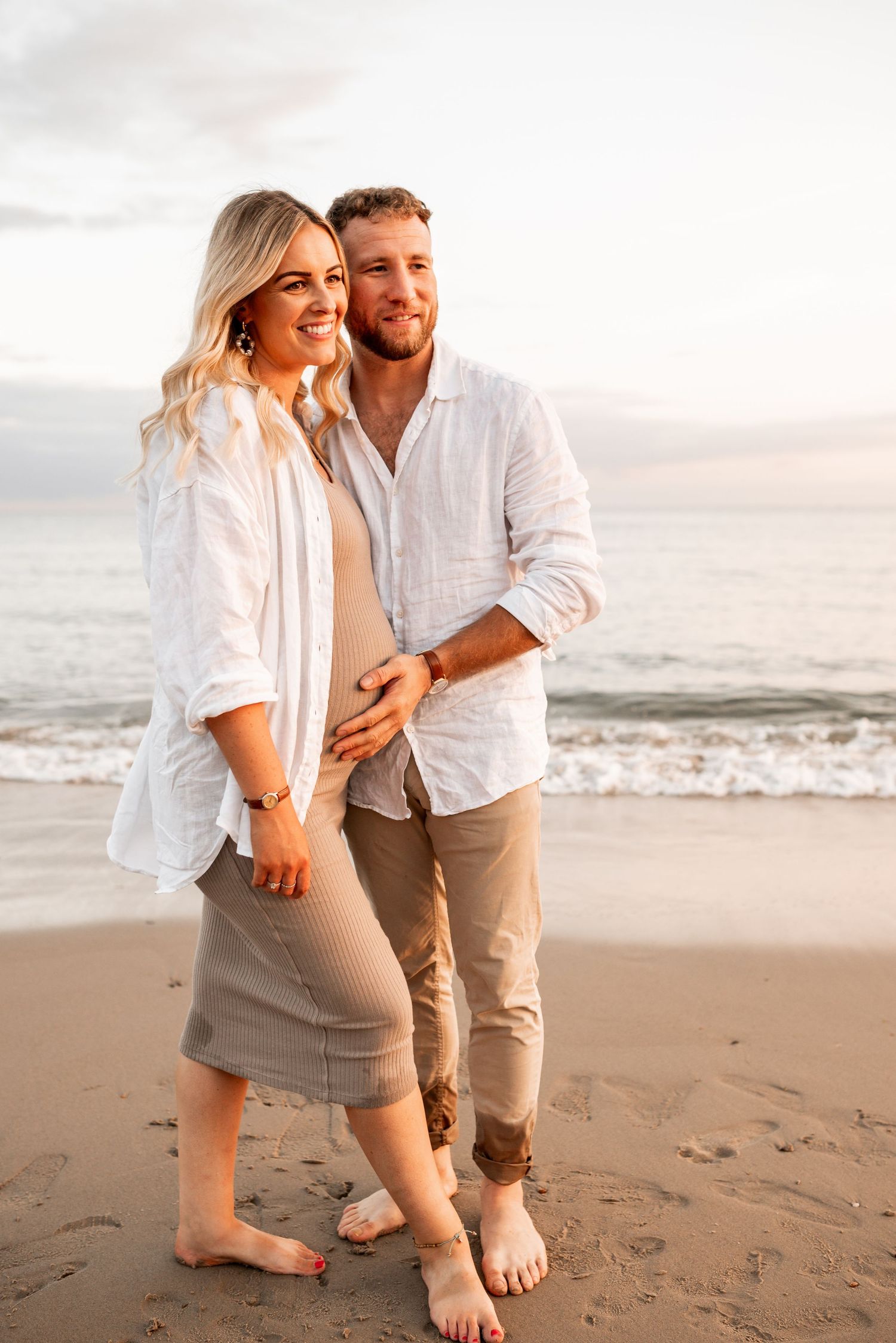 A couple in white outfits embraces on a beach at sunset with ocean waves in the background.