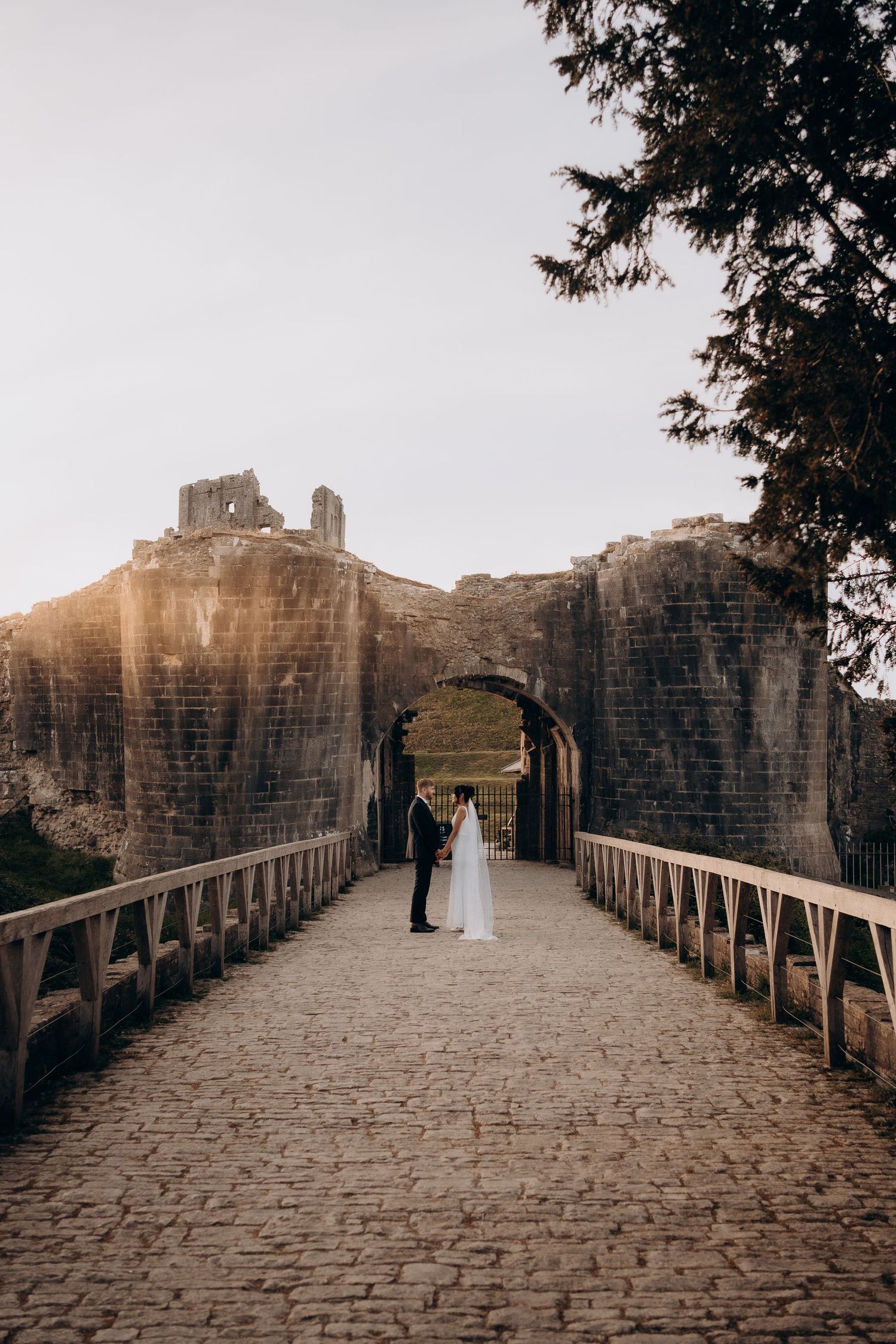 Bride and groom hold hands on cobblestone bridge leading to ancient castle ruins at golden hour.