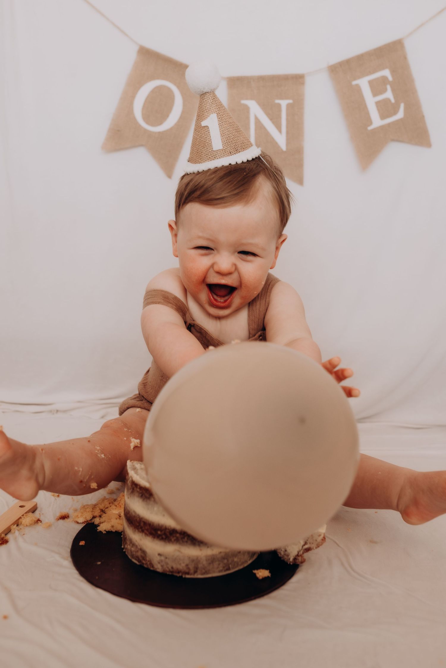 Baby in a beige sweater smiles while playing with a chocolate cake during a first birthday cake smash photo session.