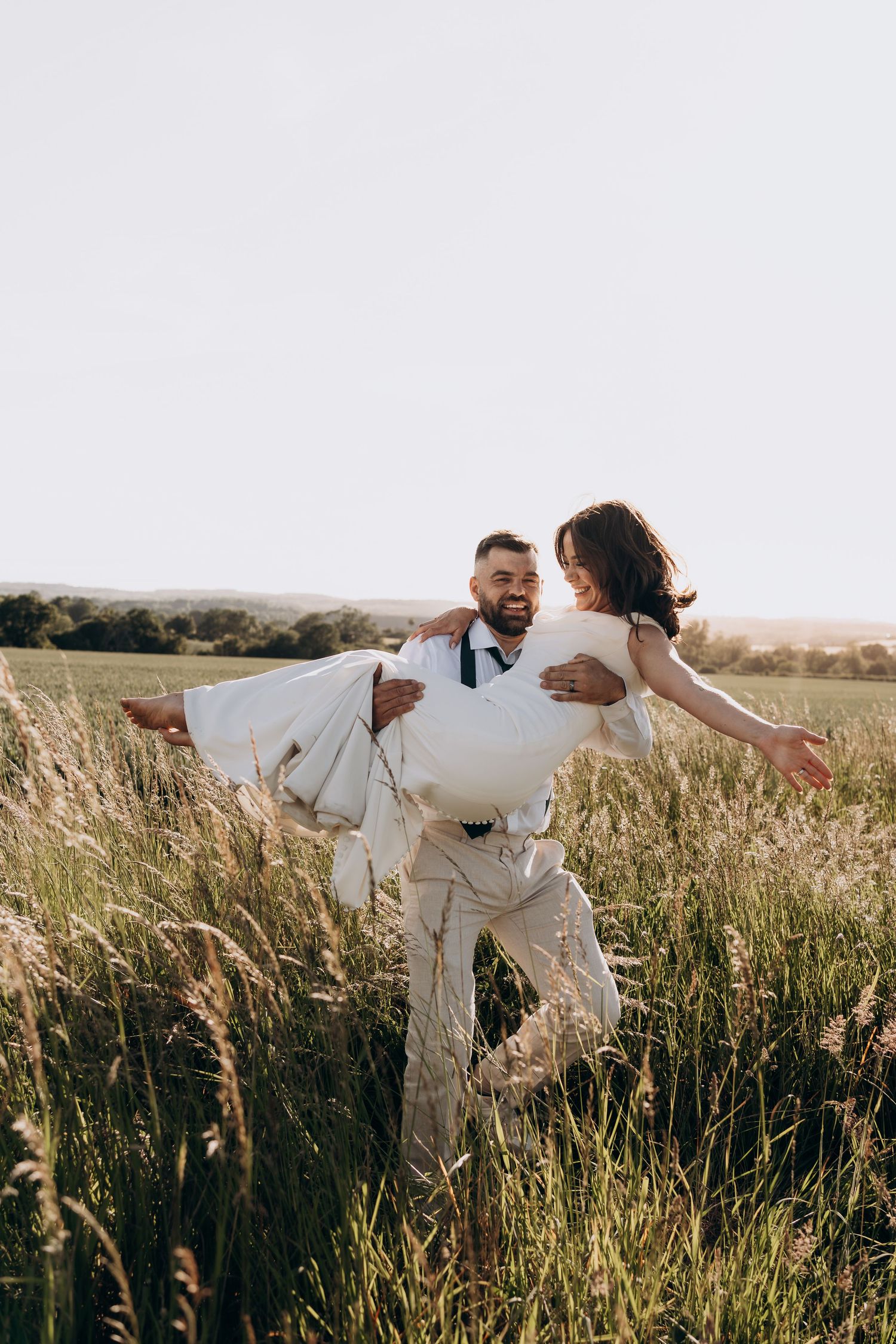 Groom carrying bride in white dress through a sunny green field, both smiling joyfully on their wedding day.