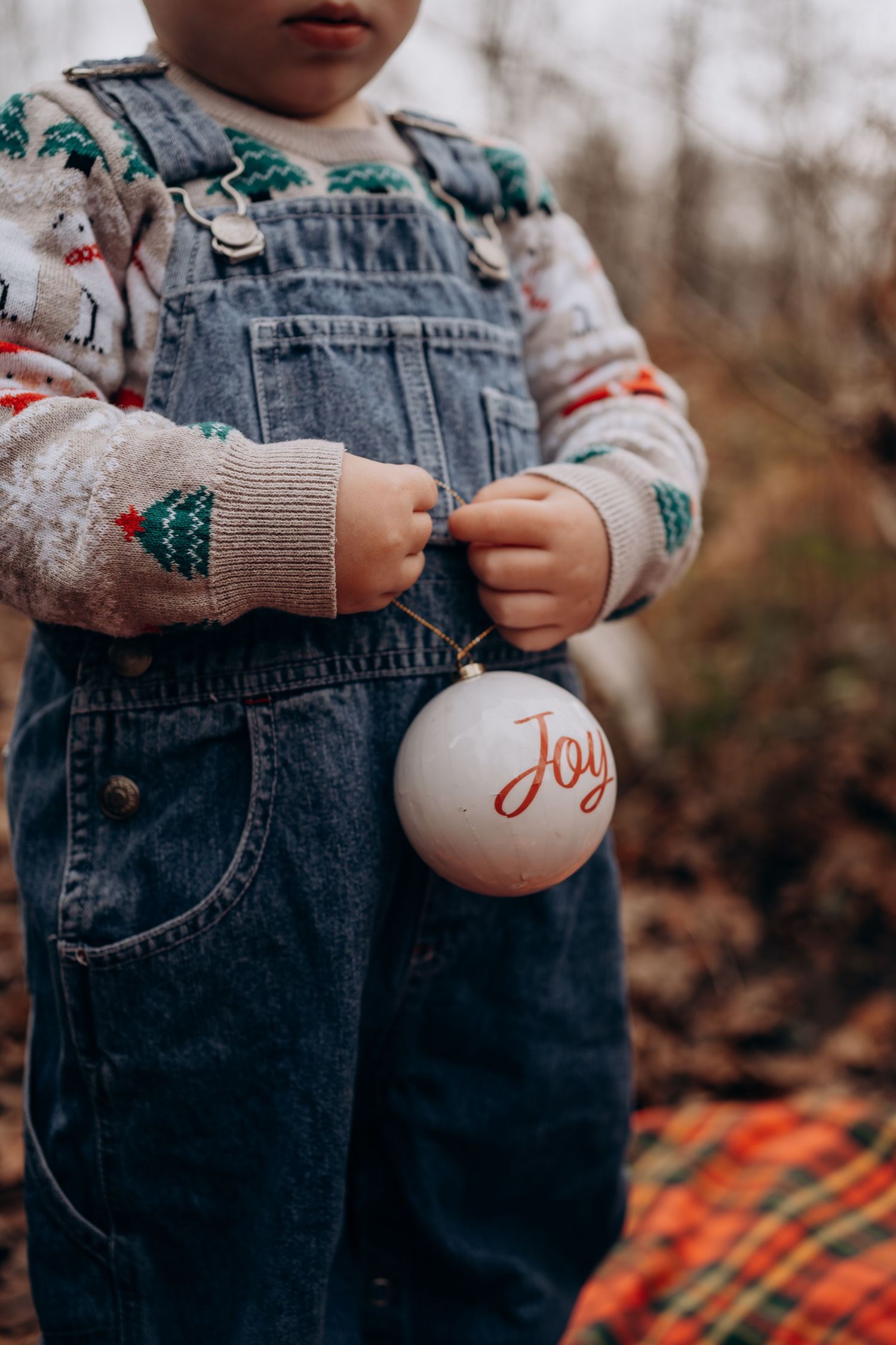 Small child in denim overalls and festive sweater holds white Christmas ornament with 'Joy' text in autumn setting.