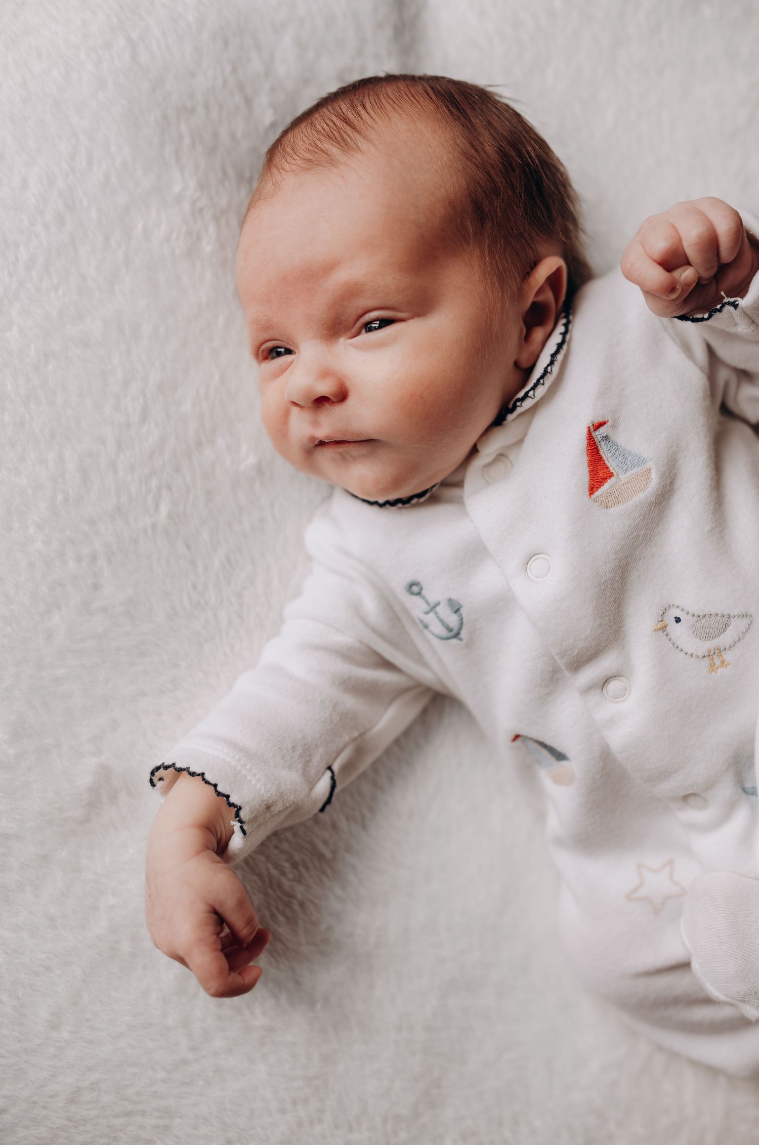 Overhead view of newborn wearing white onesie with delicate pattern lying on soft surface.