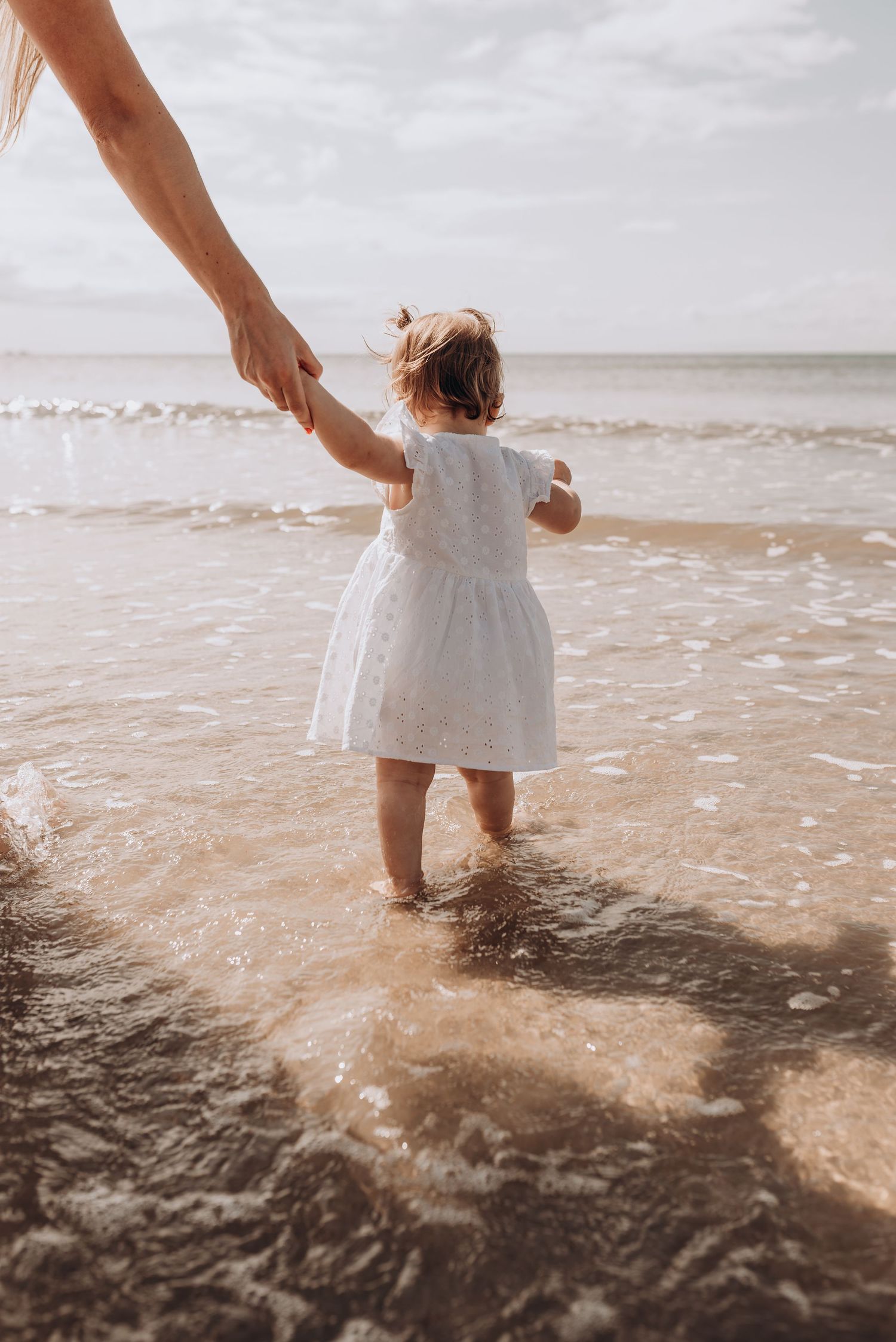 A toddler in a white dress splashes in shallow ocean waves at the beach.
