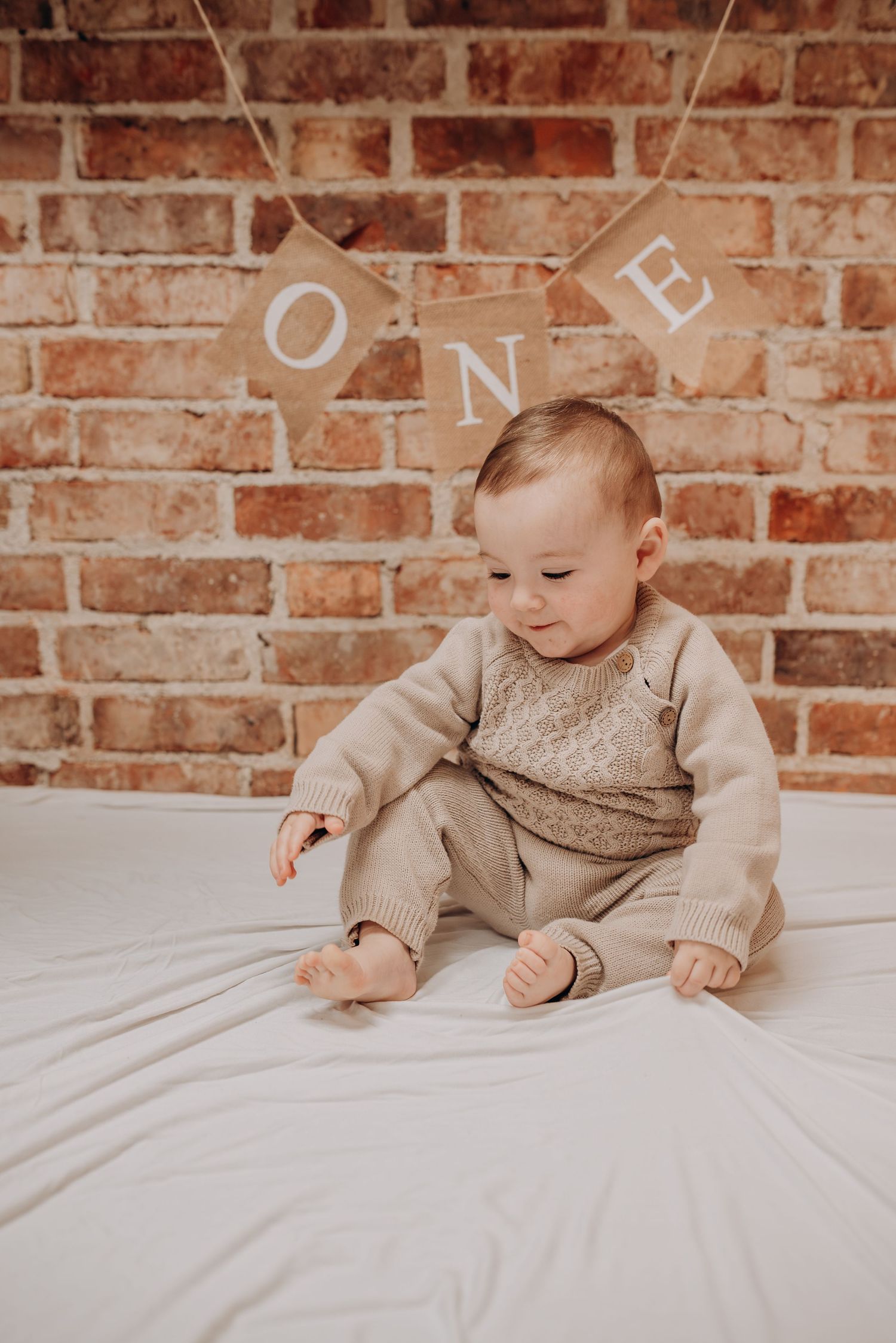 Baby in beige outfit crawls on white bedding against brick wall backdrop.