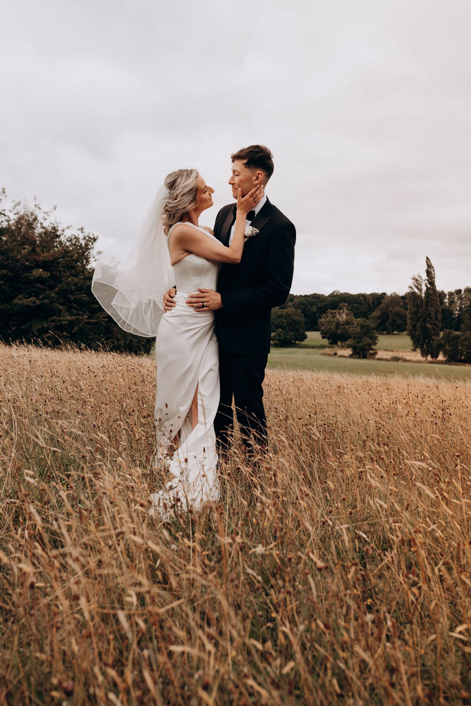 A romantic couple embraces in a golden wheat field during a wedding photoshoot under cloudy skies.