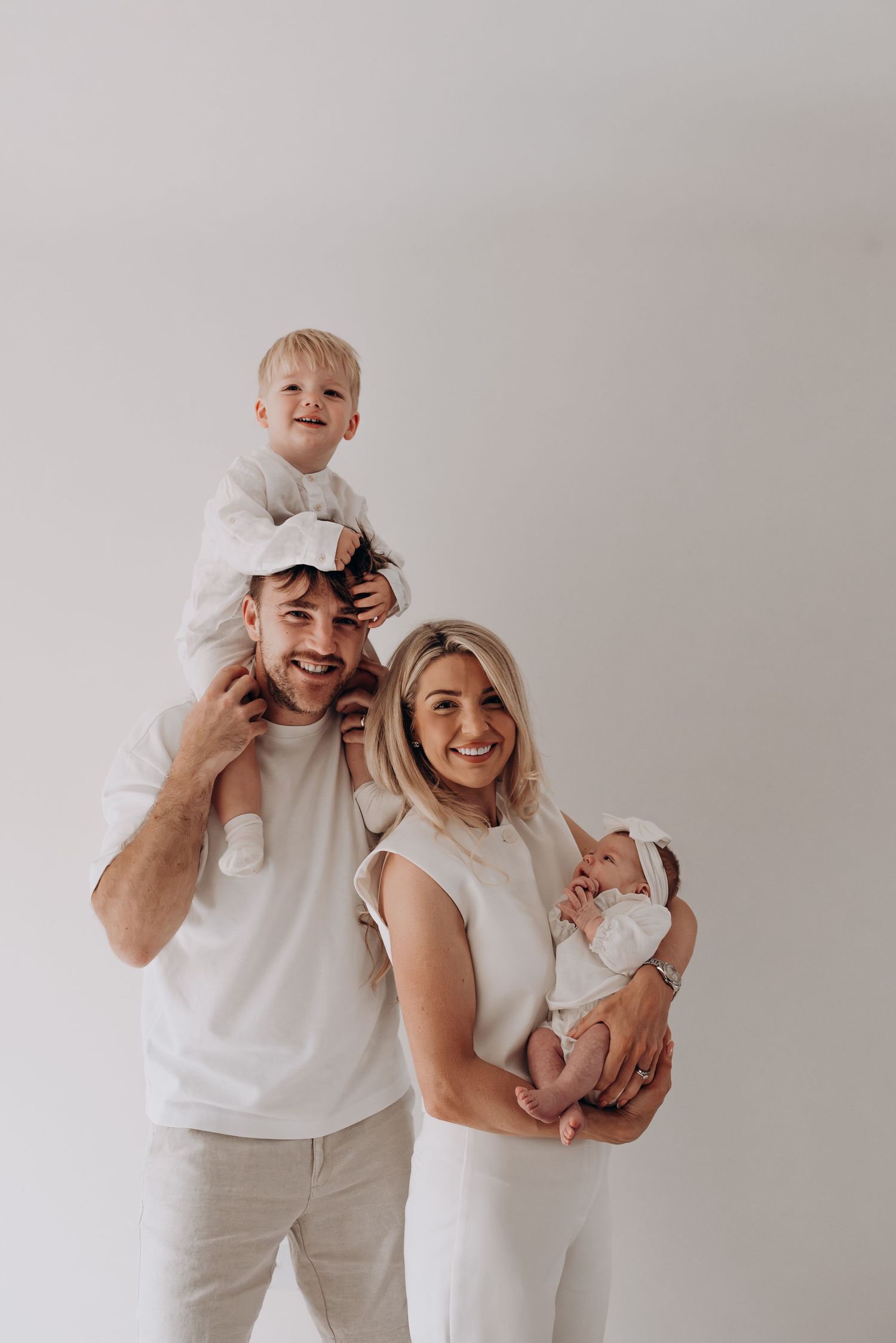 Family wearing white and cream outfits poses together against neutral background.