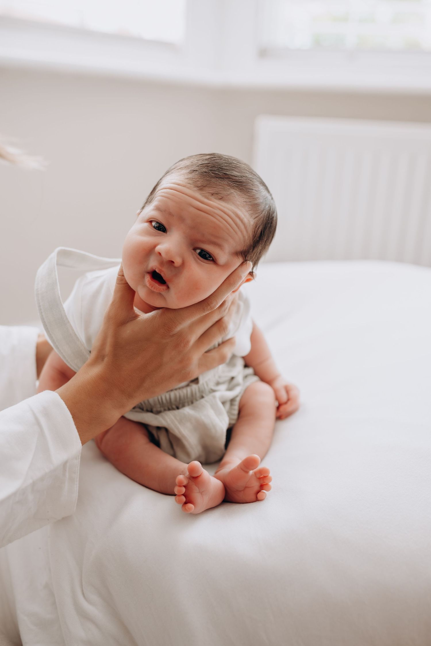 Baby in white outfit lying on white bedding with expressive facial gestures.