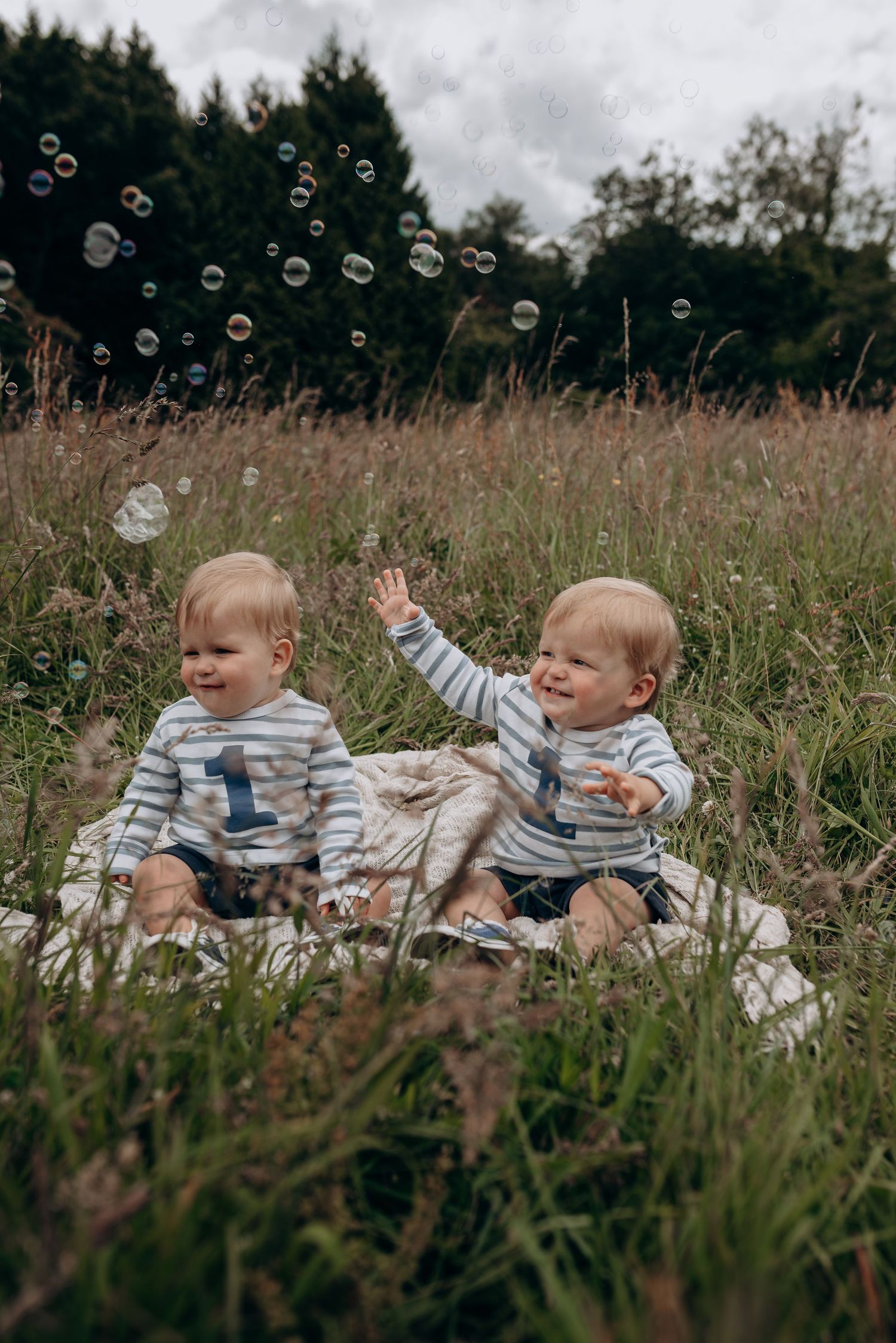 Two babies in striped shirts play with bubbles in a grassy field.