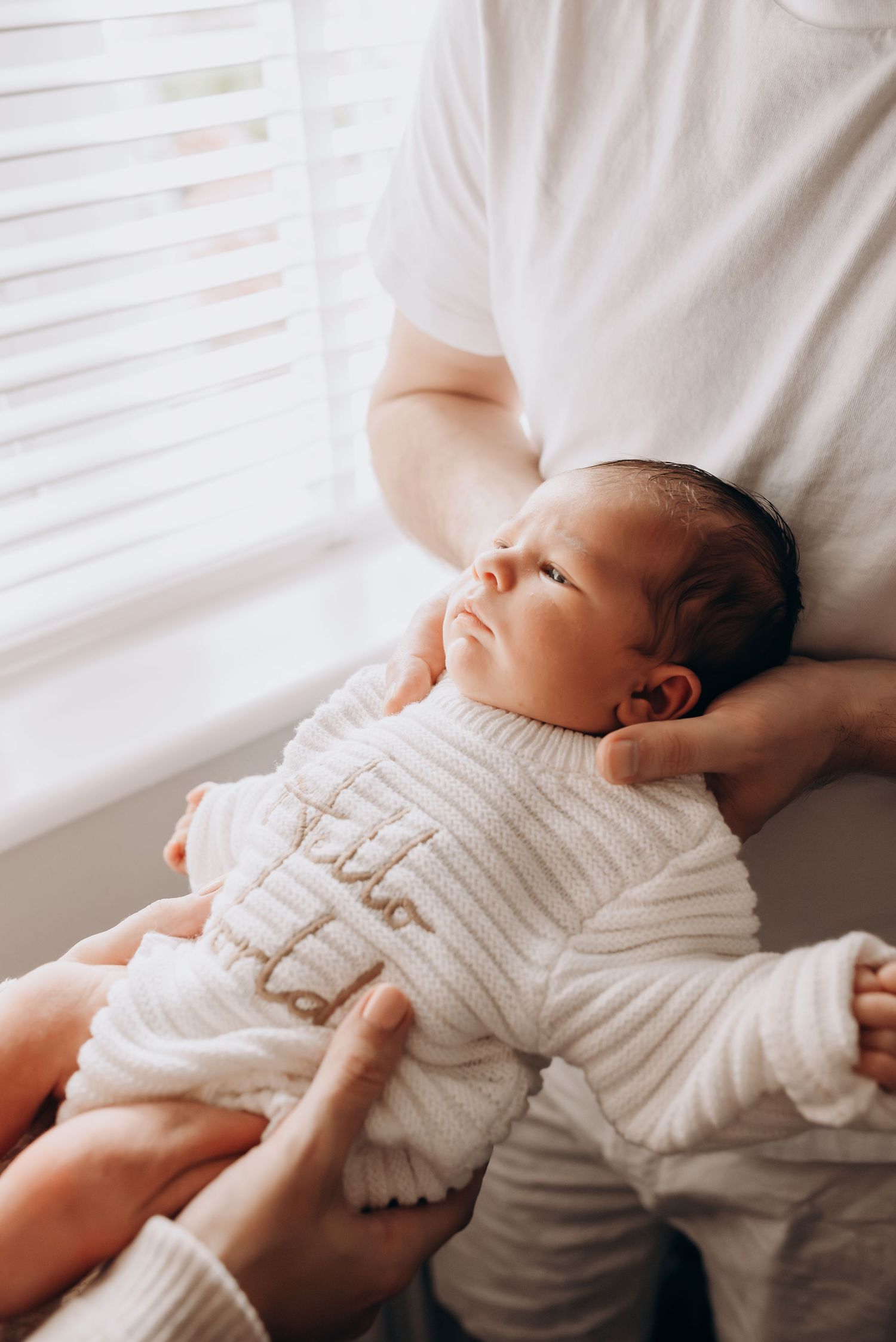 Newborn baby in white knit sweater resting peacefully near window light.