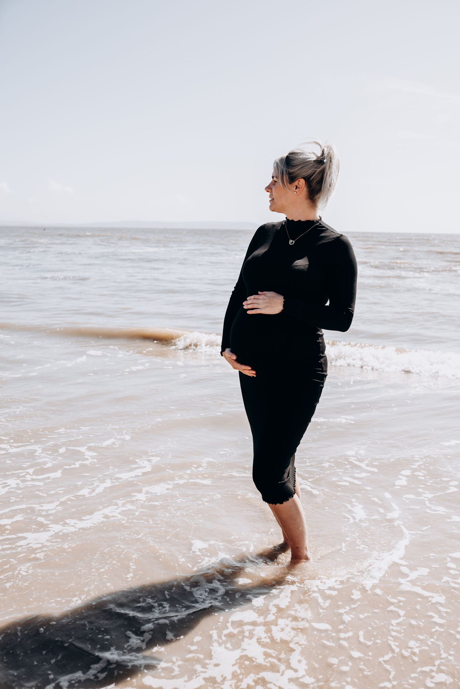 Someone walks through gentle ocean waves wearing dark clothing on a bright beach.