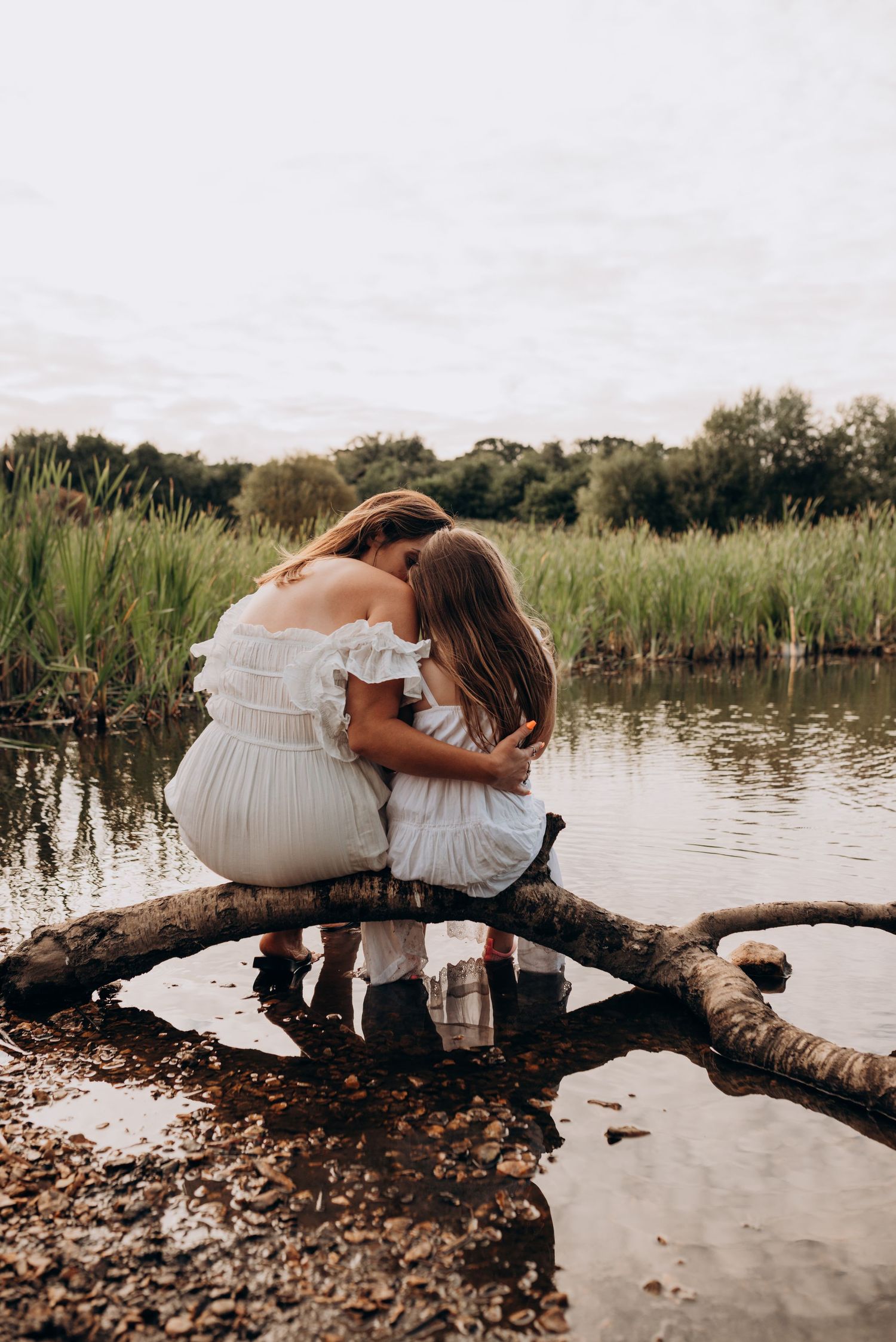 Two people in white dresses sit together on a fallen log over a serene lake at sunset.