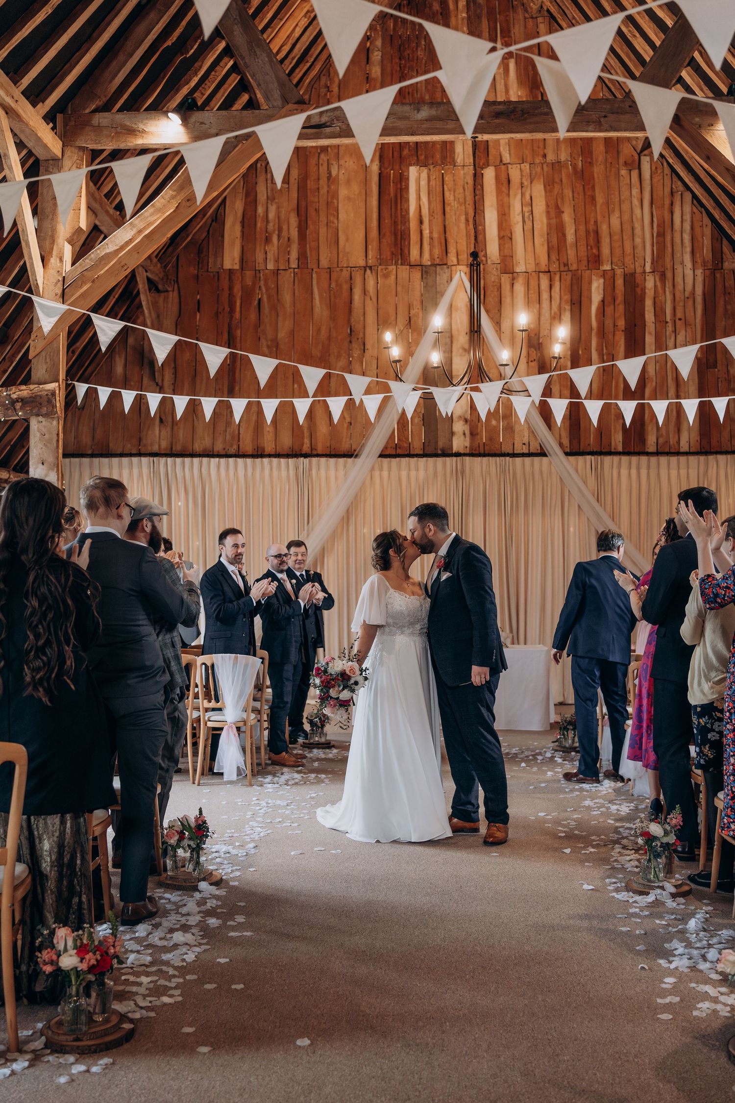 Rustic barn wedding ceremony with white bunting decorations and guests gathered in a warm wooden interior.