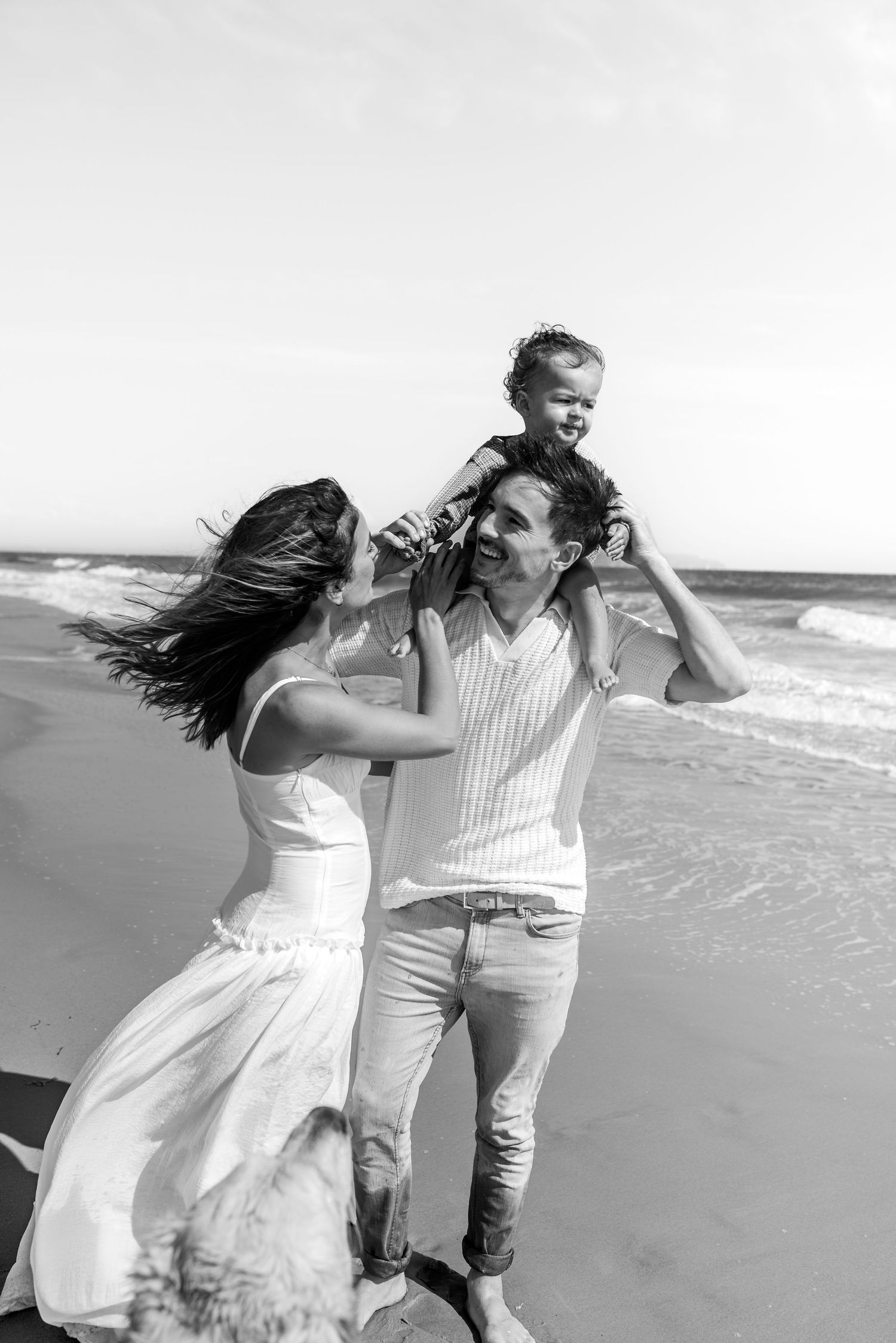 Romantic couple walking along a windy beach with flowing white dress and casual attire.