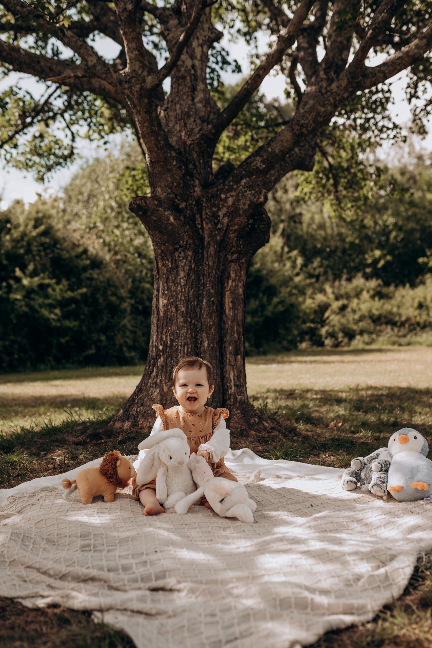 Baby lying on white blanket under large tree in outdoor setting.