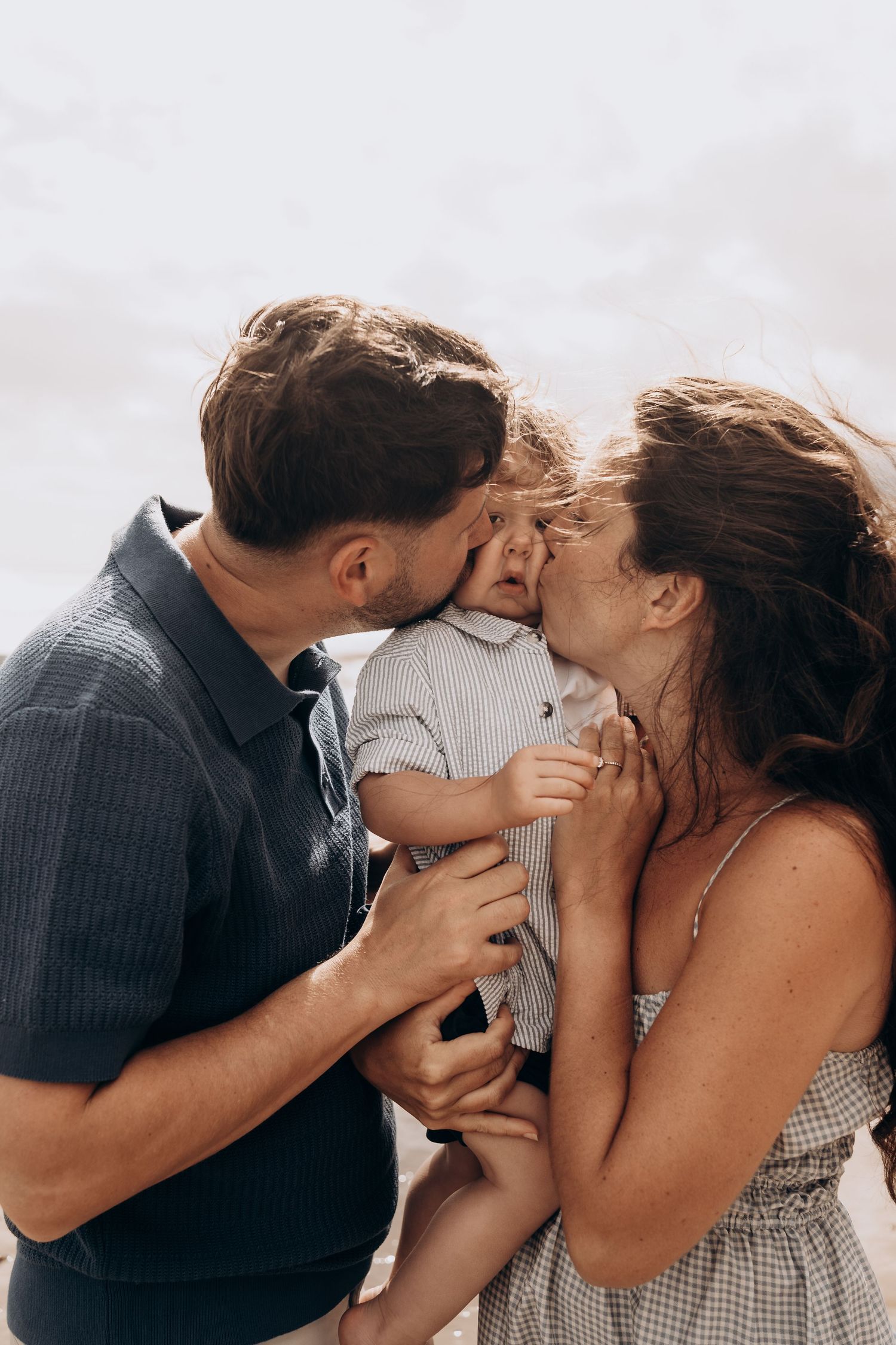 Parents kissing their baby on the cheeks outdoors, a tender family moment captured at the beach.