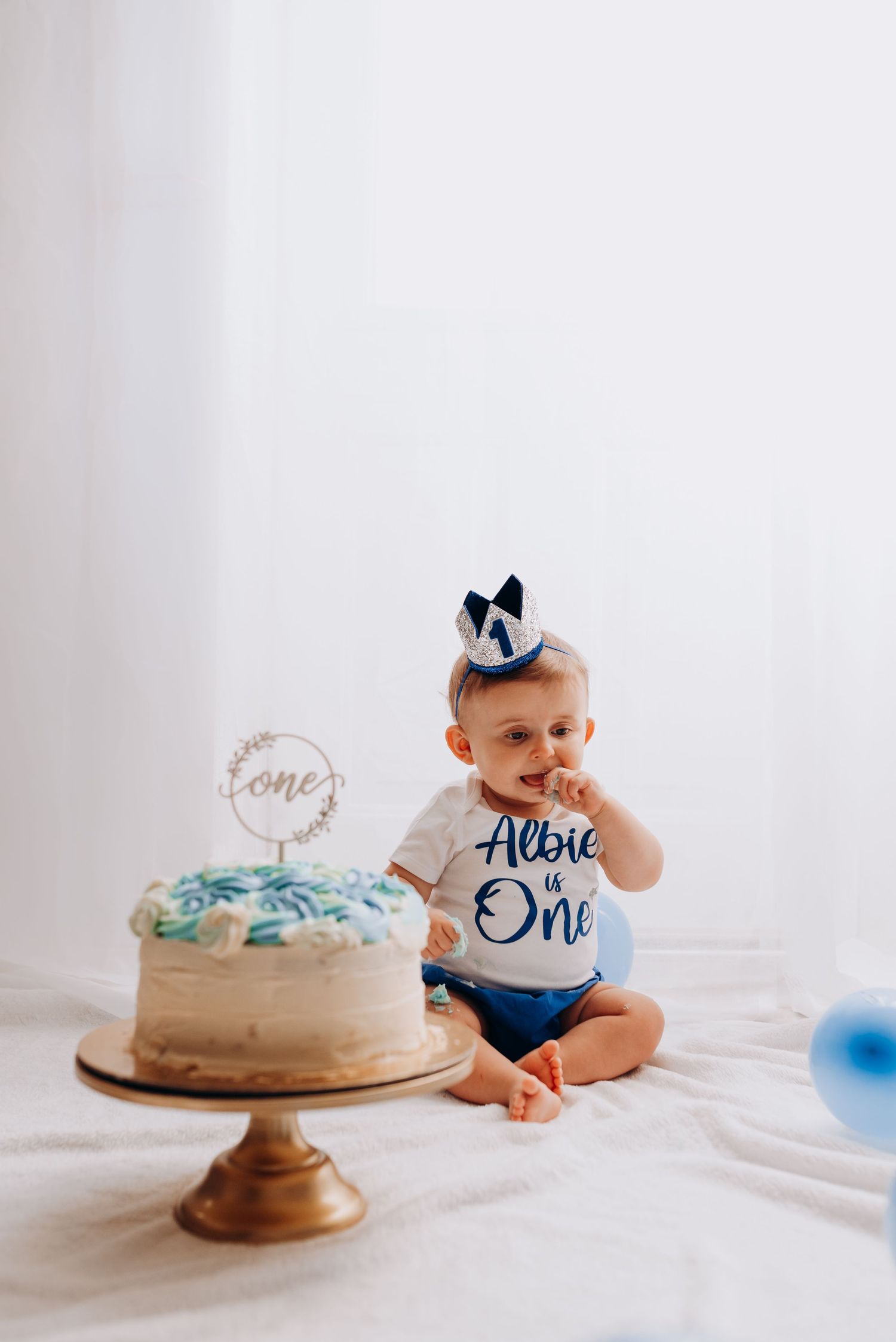First birthday cake smash setup with blue themed decorations and baby reaching for frosted cake.