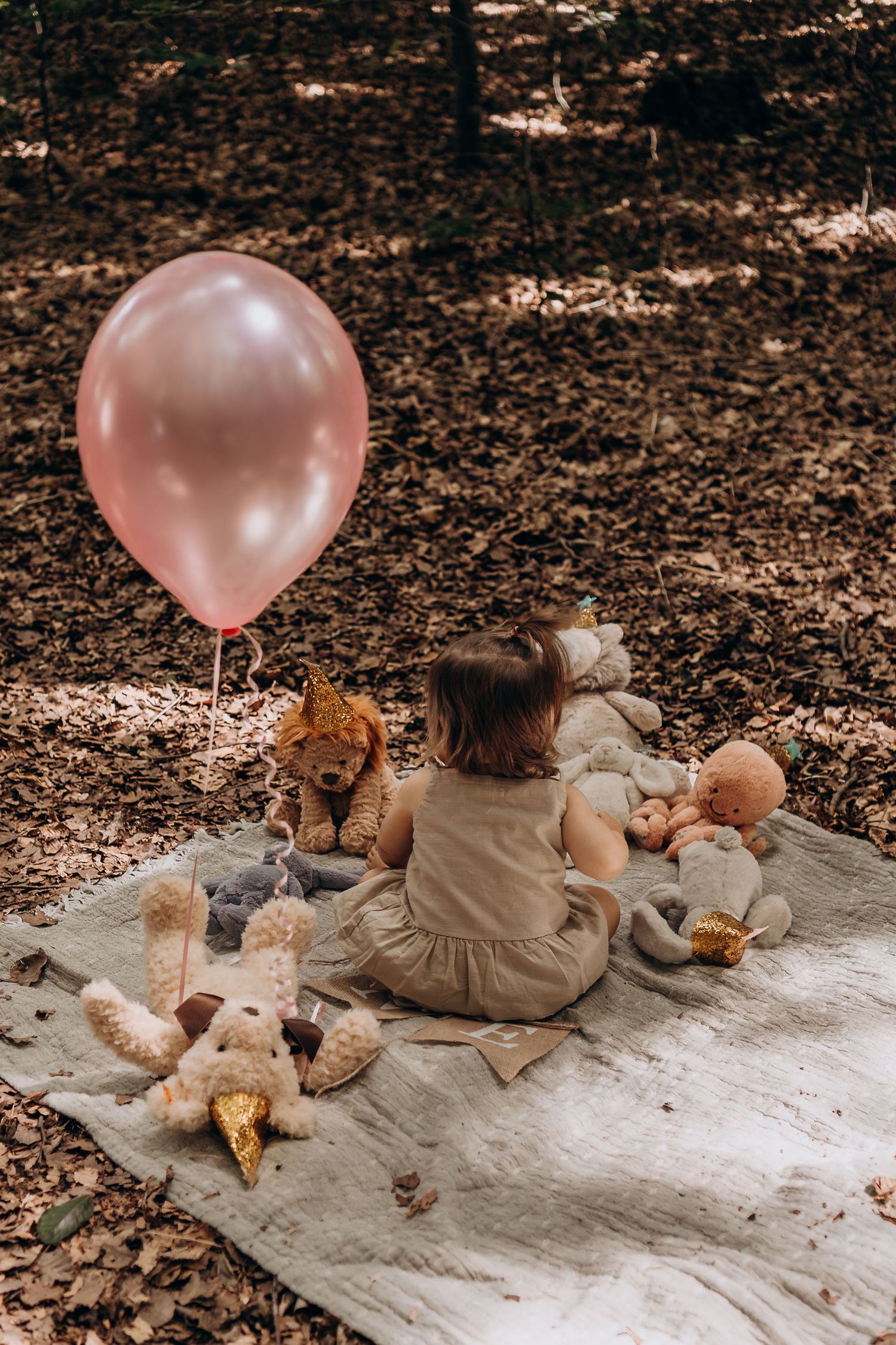 A child sits on a blanket in autumn leaves with stuffed animals and a pink balloon.