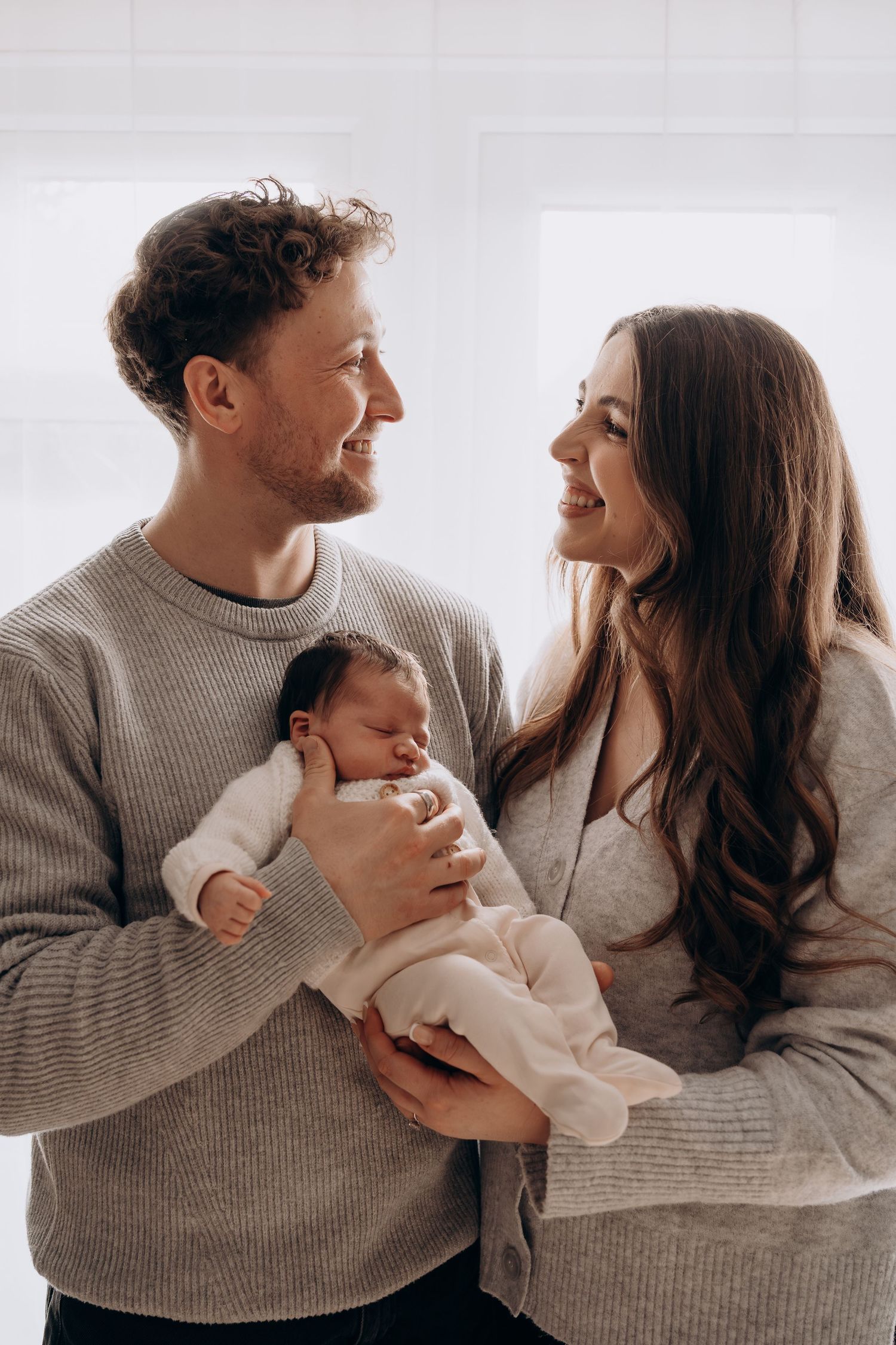 A couple in matching gray sweaters share tender moments while holding their baby in a series of intimate photos.