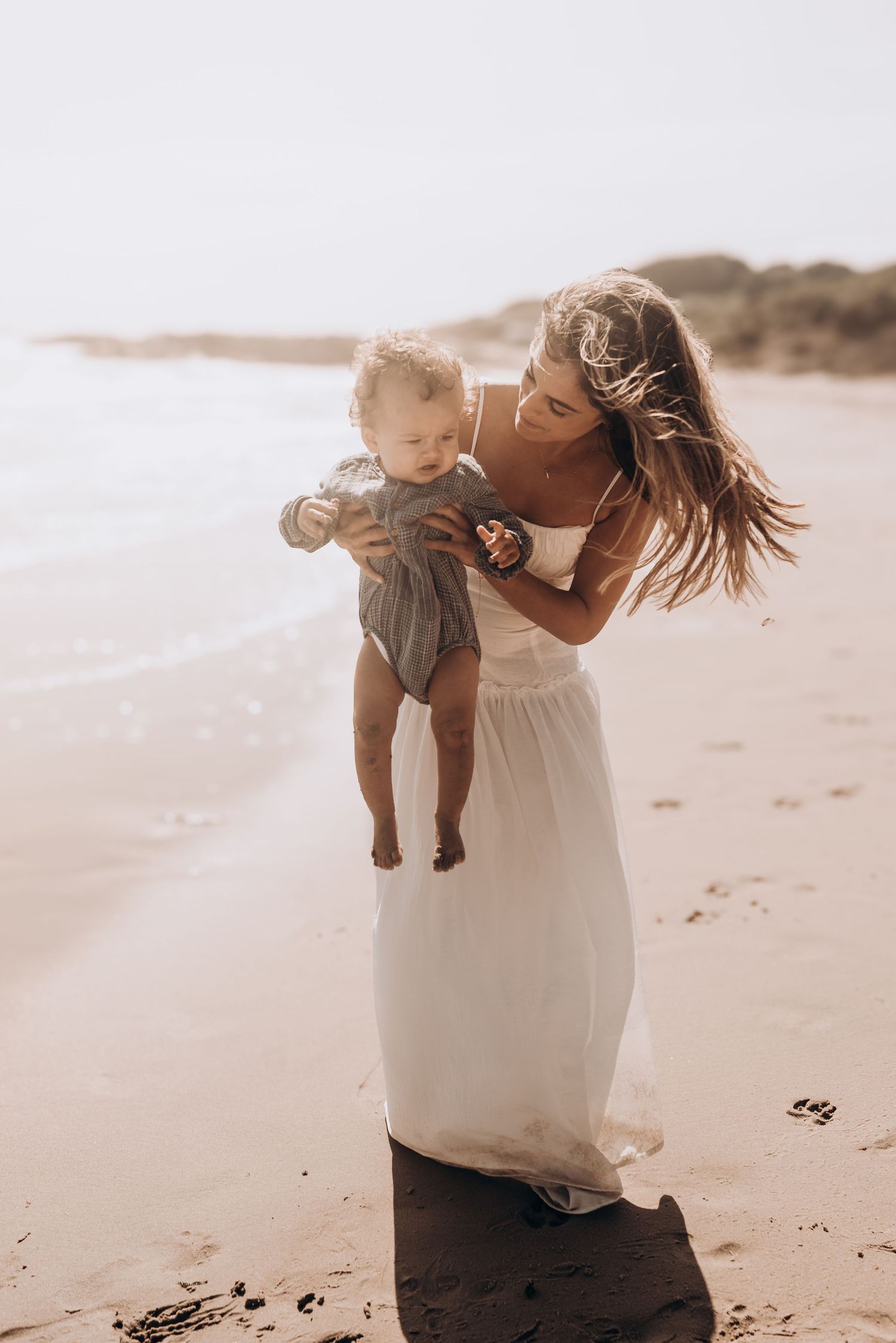Mother in white dress playing with baby on sunny beach.