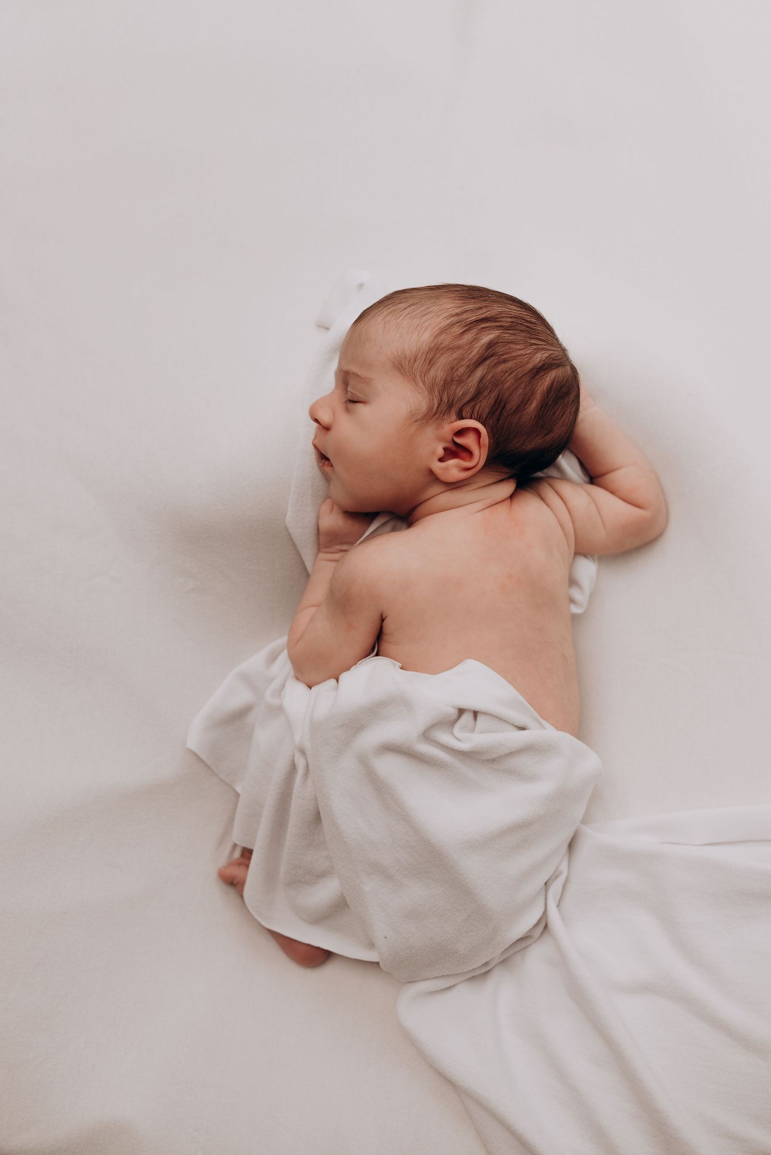 Sleeping newborn baby in white onesie rests on light colored bedding.