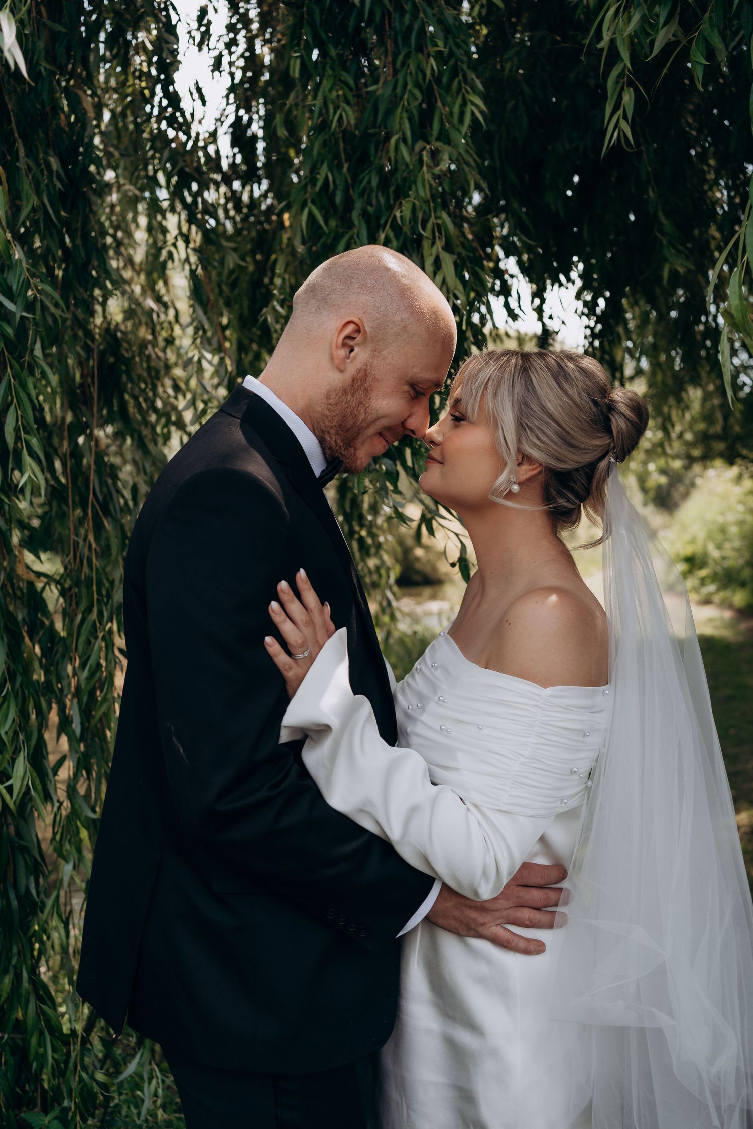Bride and groom share a romantic moment under weeping willow, bride in off-shoulder gown with veil, groom in black tuxedo.