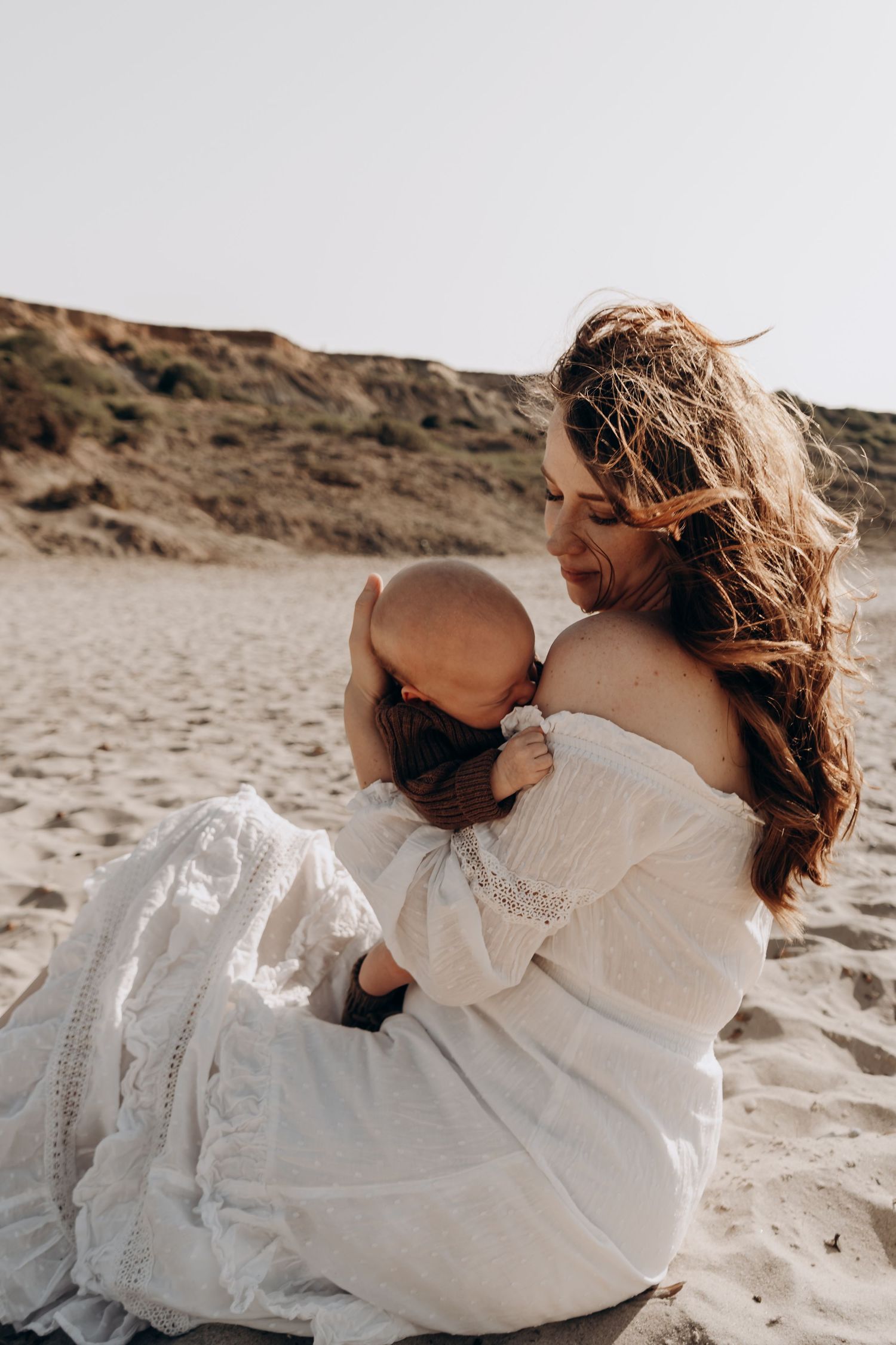 A mother in a white off-shoulder dress breastfeeding her baby on a sandy beach with dunes in the background.