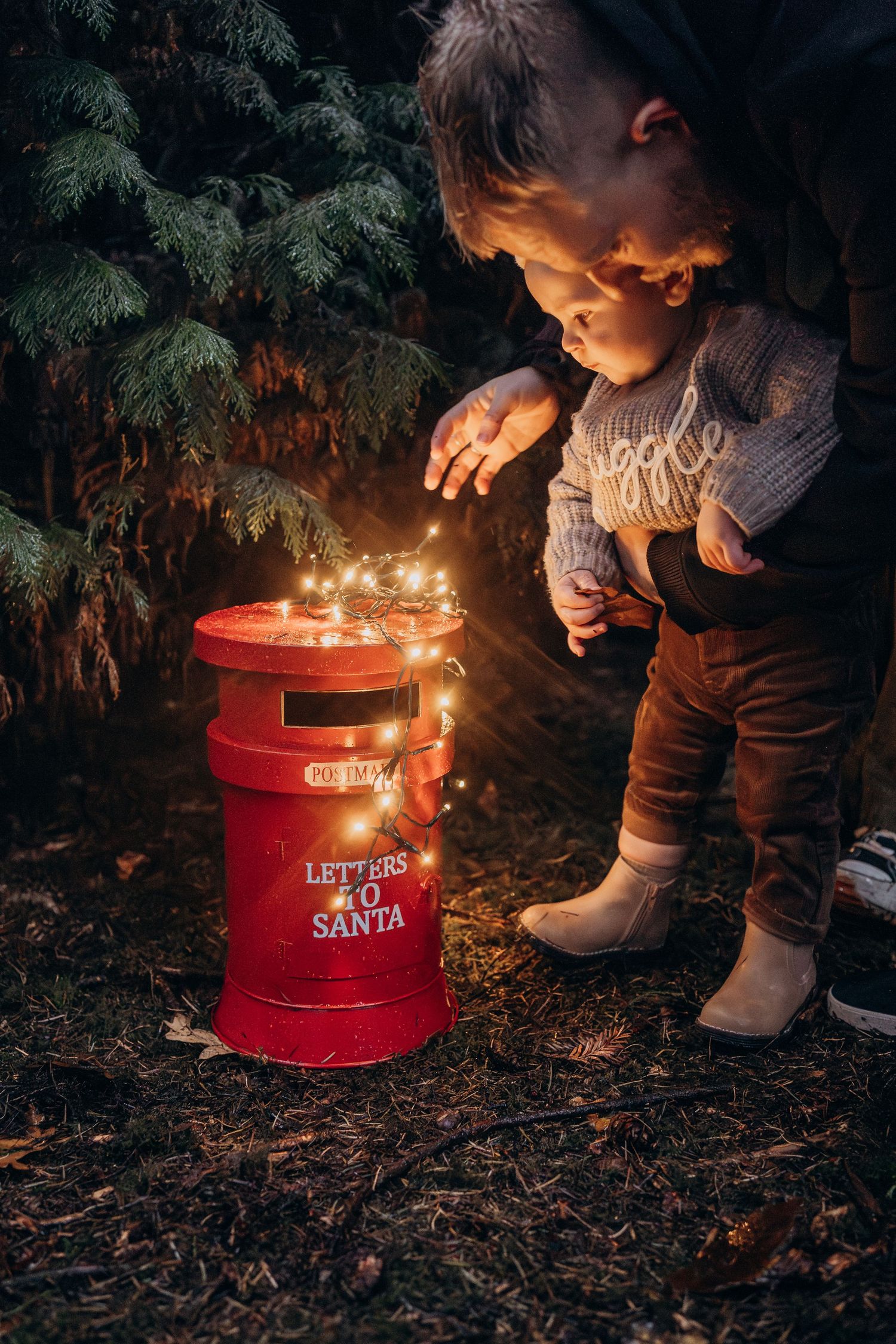A red Santa's mailbox illuminated by candlelight glows warmly against a dark evergreen background at night.