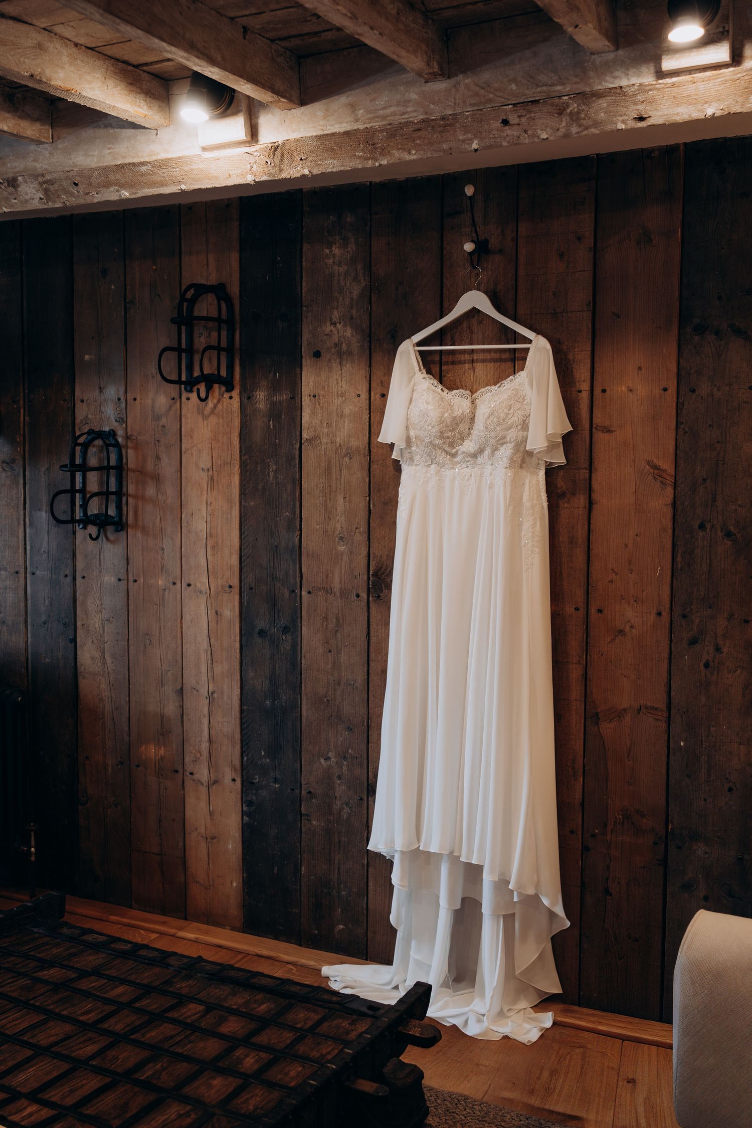 A white wedding dress hangs on a wooden wall in a rustic room with exposed beams and warm lighting.