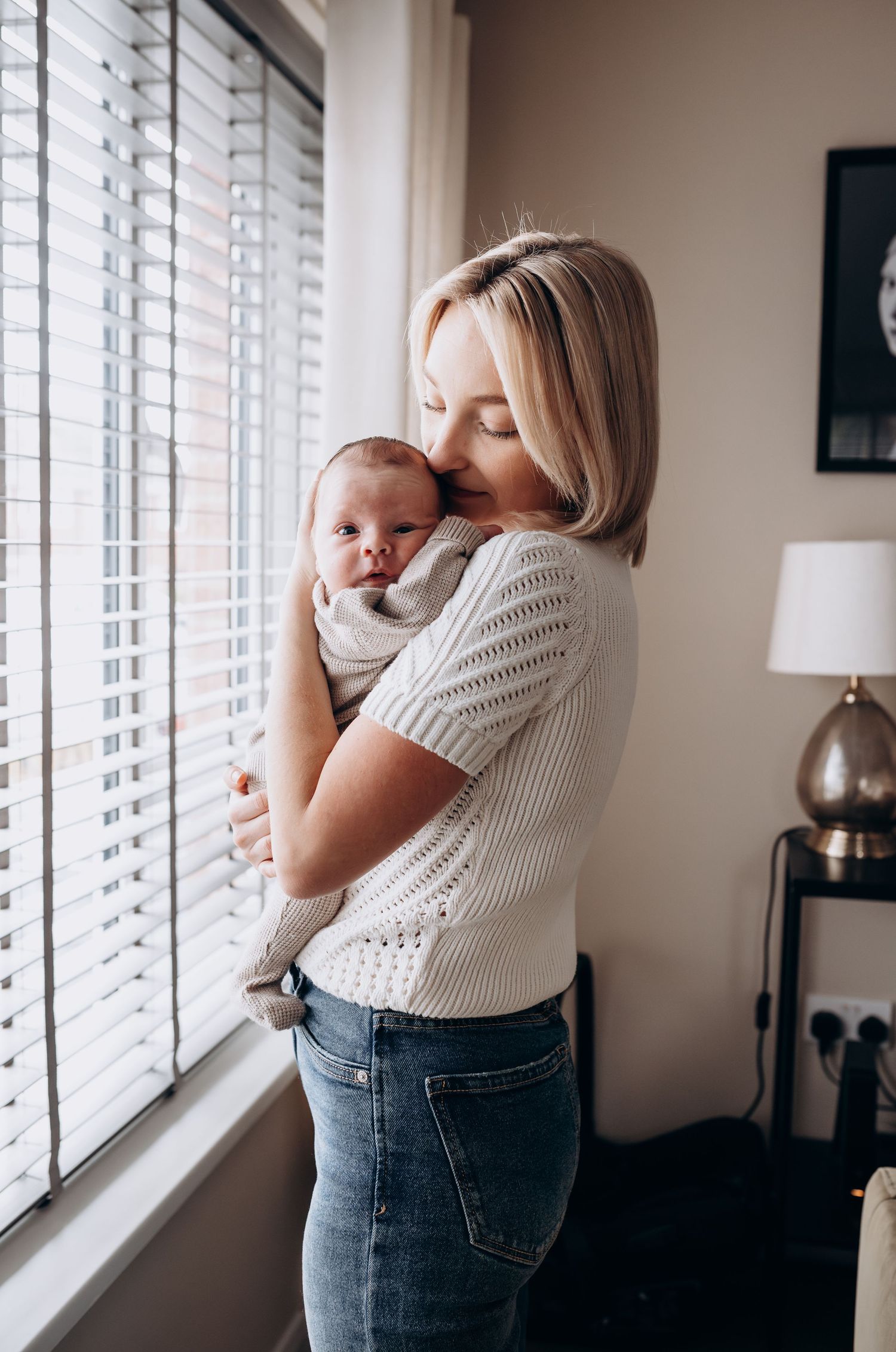 Parent and young child share tender moment by window with natural light streaming through white blinds.