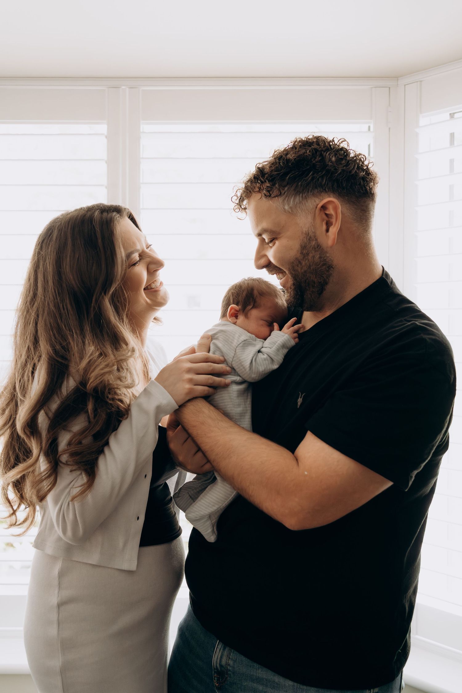 Happy couple smiling at their newborn baby by a bright window in a cozy home setting.