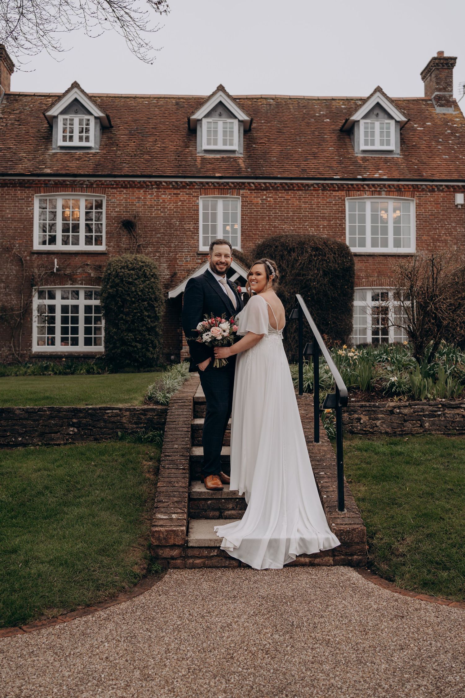 A couple shares a romantic moment outside a traditional English brick manor house with gravel driveway and manicured gardens.