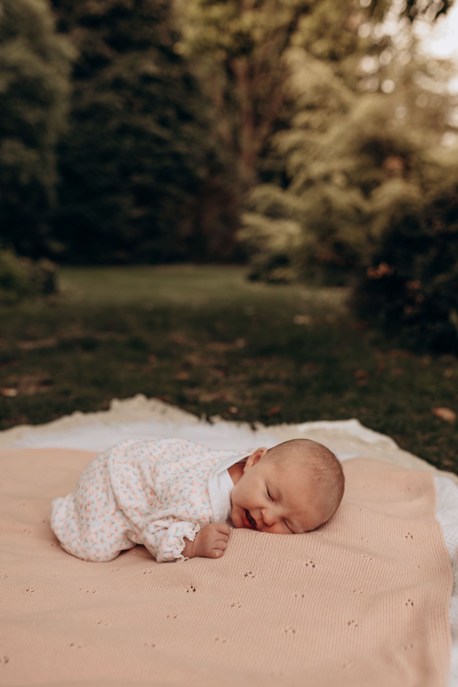 Sleeping newborn baby in white outfit on peach blanket outdoors.
