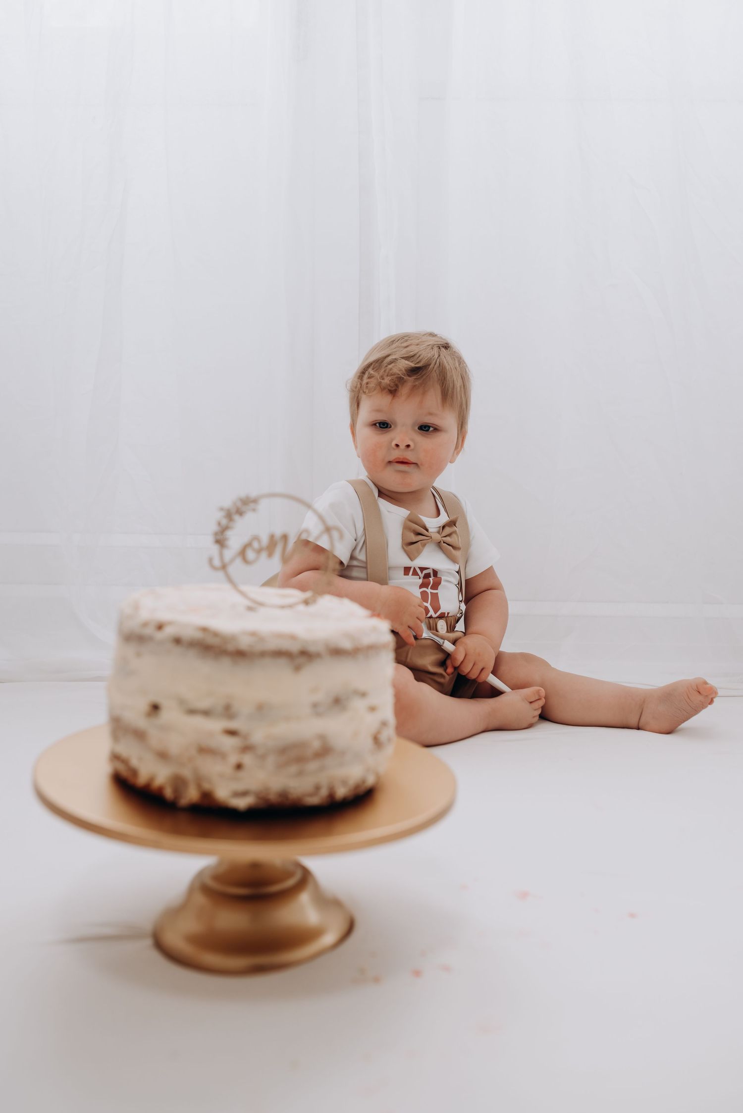 White cake on gold stand with minimalist decorations against white background.