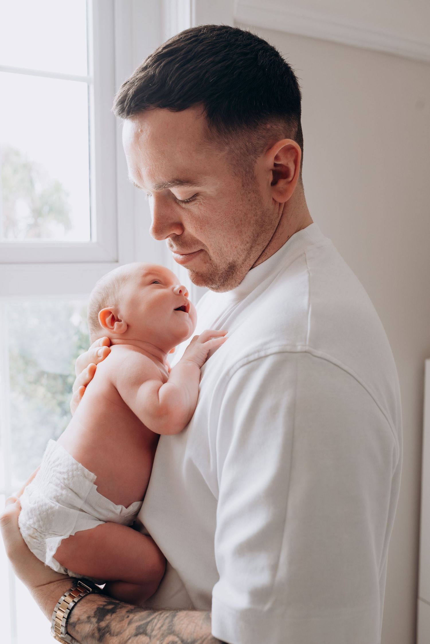 Parent in white shirt tenderly holding sleeping newborn baby near window.