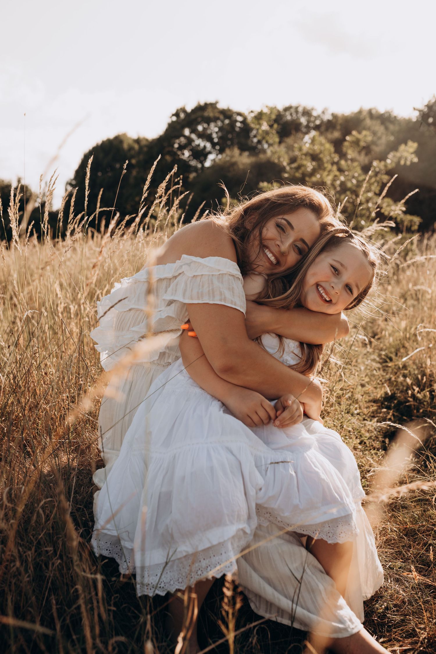 Two people in white dresses embrace while sitting in a golden sunlit field of tall grass at sunset.