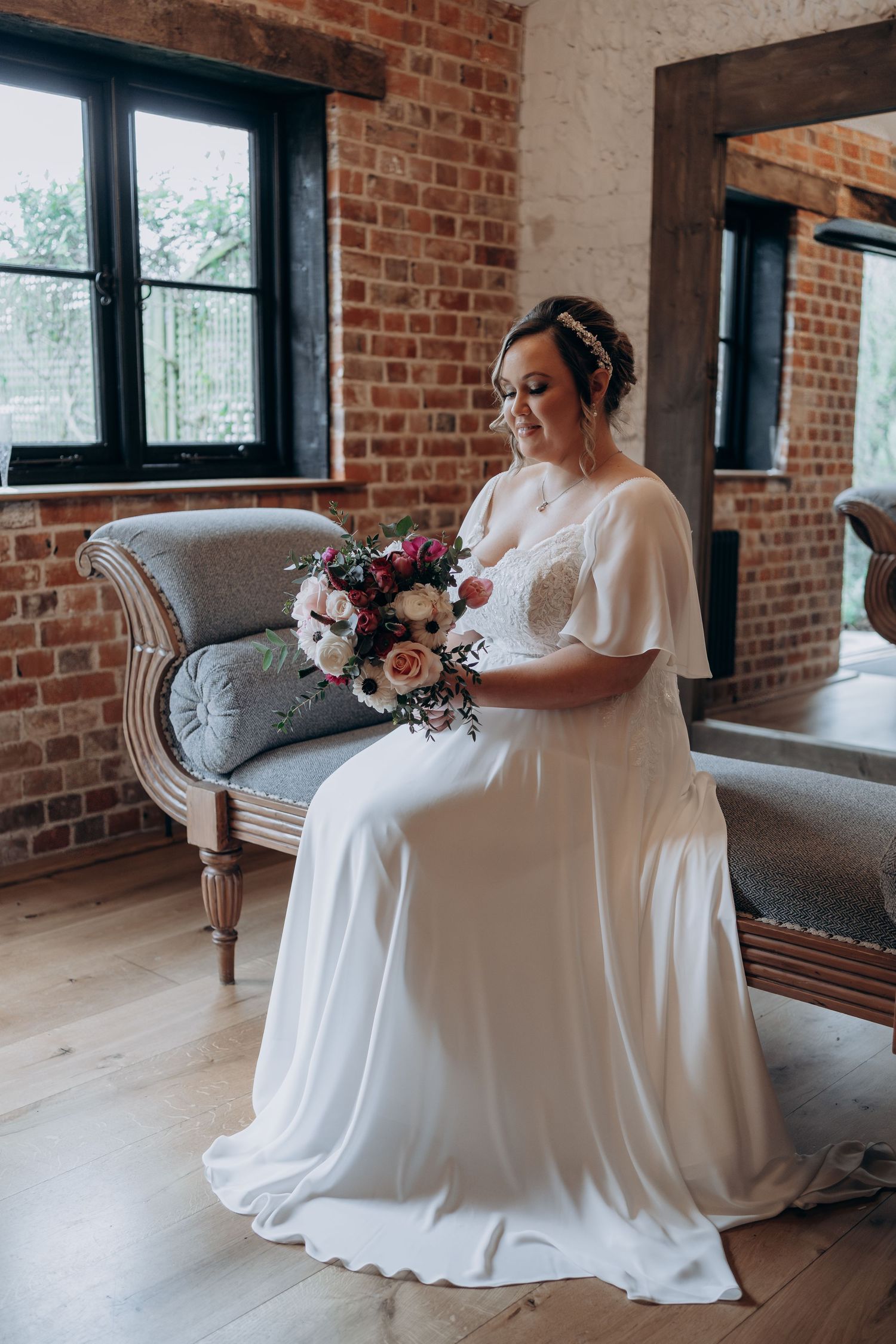 A bride in a flowing white wedding dress sits gracefully on a vintage chair against exposed brick walls.