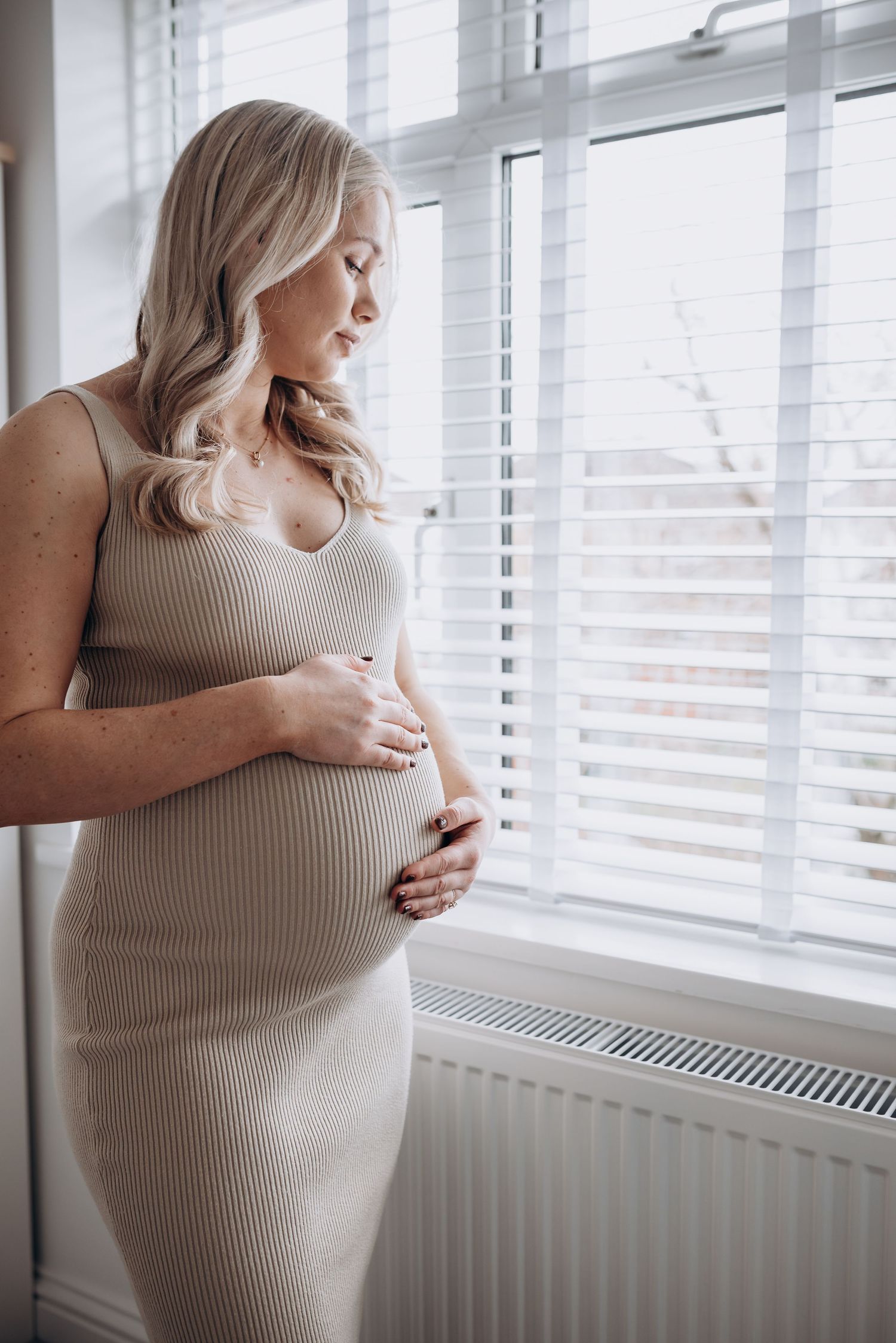 A person in a ribbed dress stands by a window, cradling their pregnant belly.