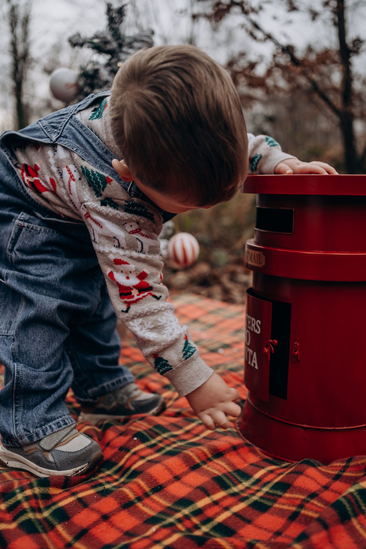 Small child reaches into vintage red mailbox on plaid blanket during autumn outdoor scene.