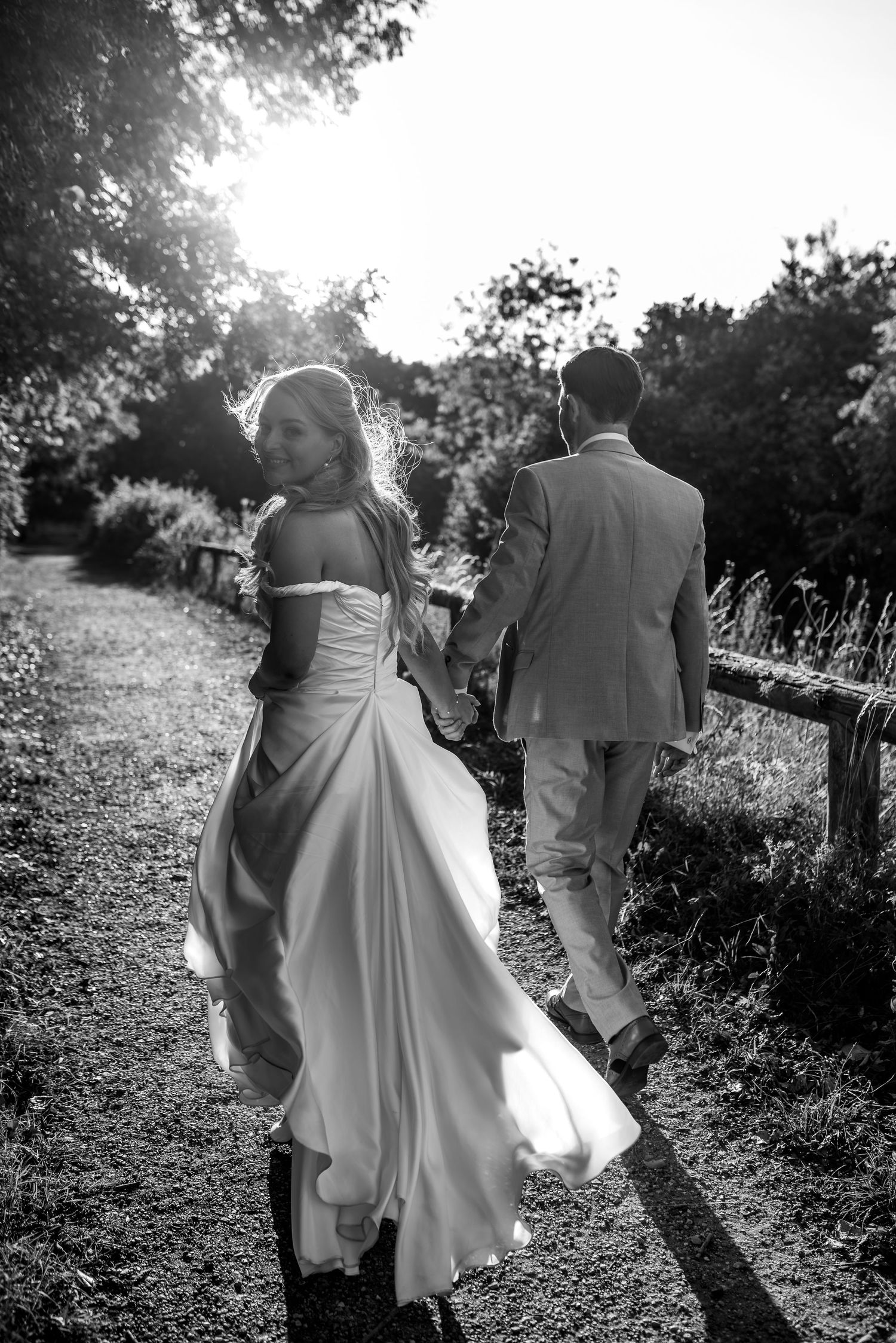 A bride in a flowing white dress and groom walking along a sunlit garden path in black and white photography.