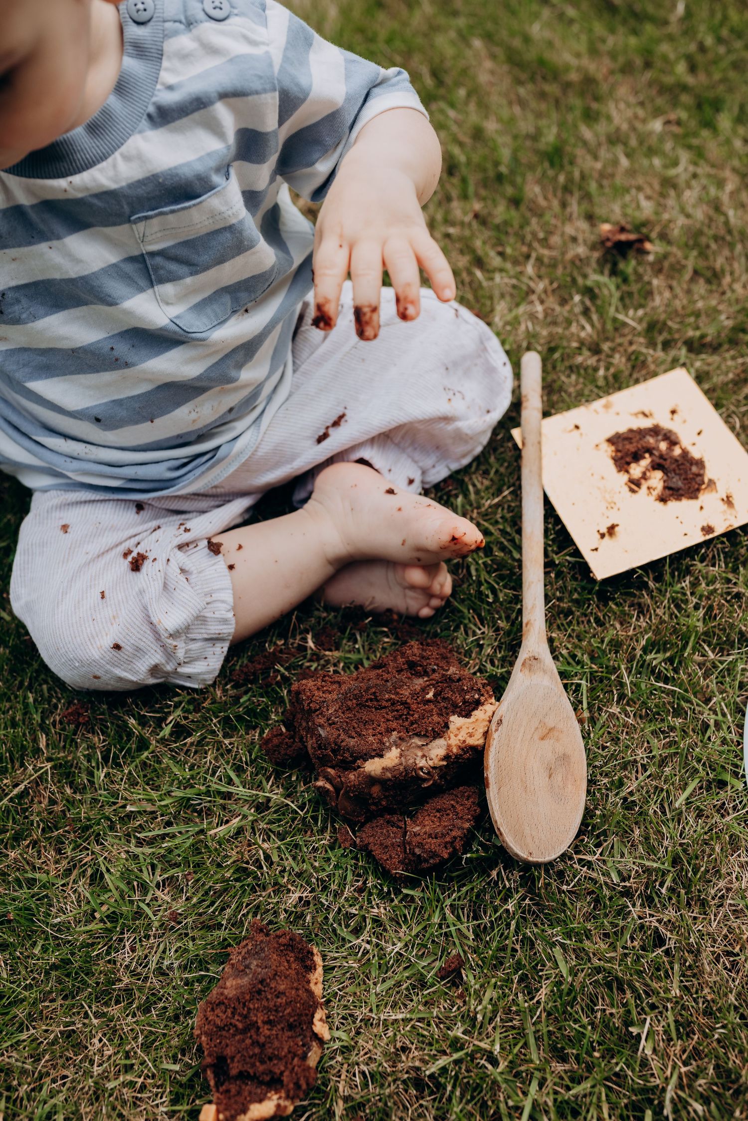Small hands playing with chocolate cake and wooden spoon on grass.