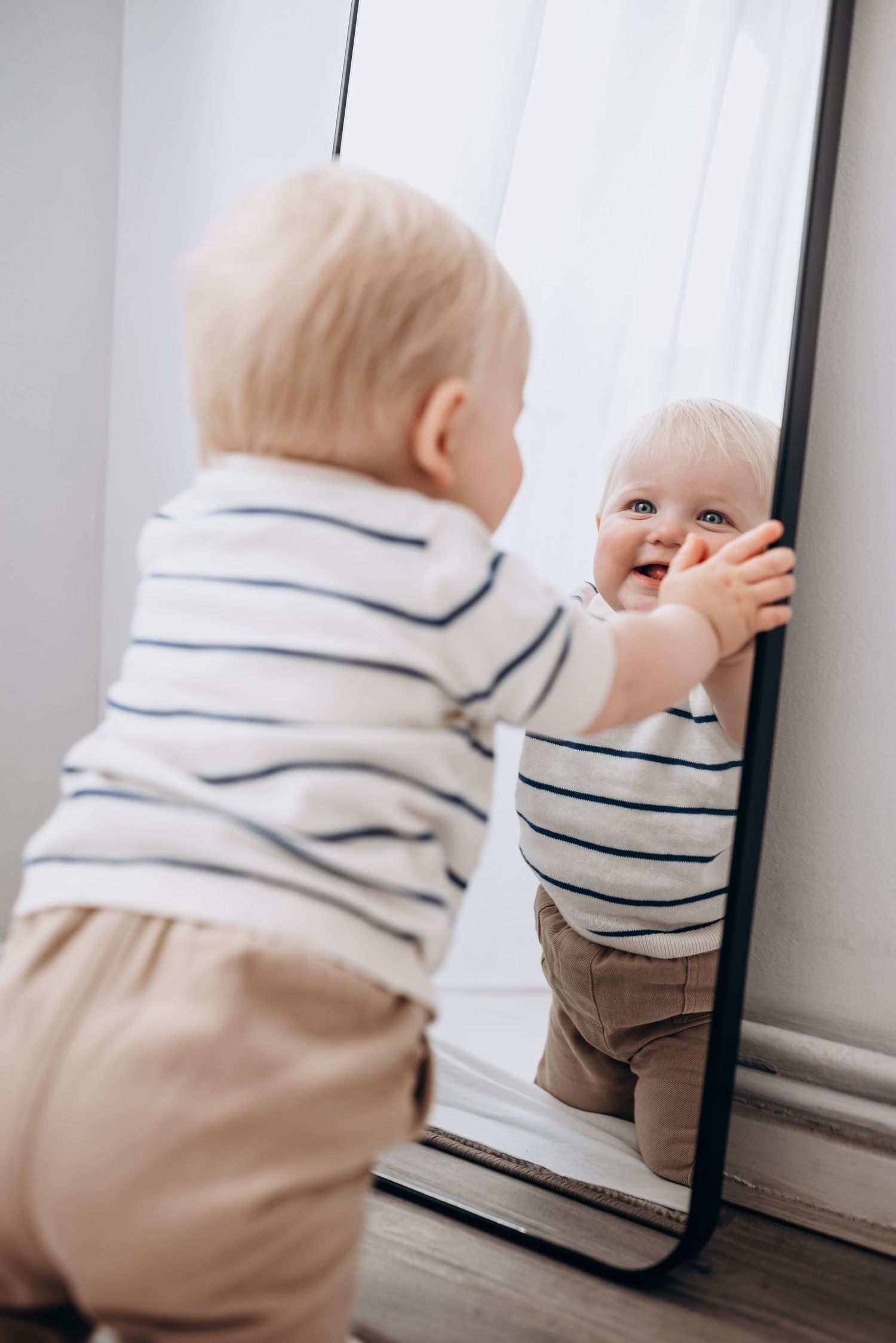 Baby in striped outfit crawling towards mirror on hardwood floor.