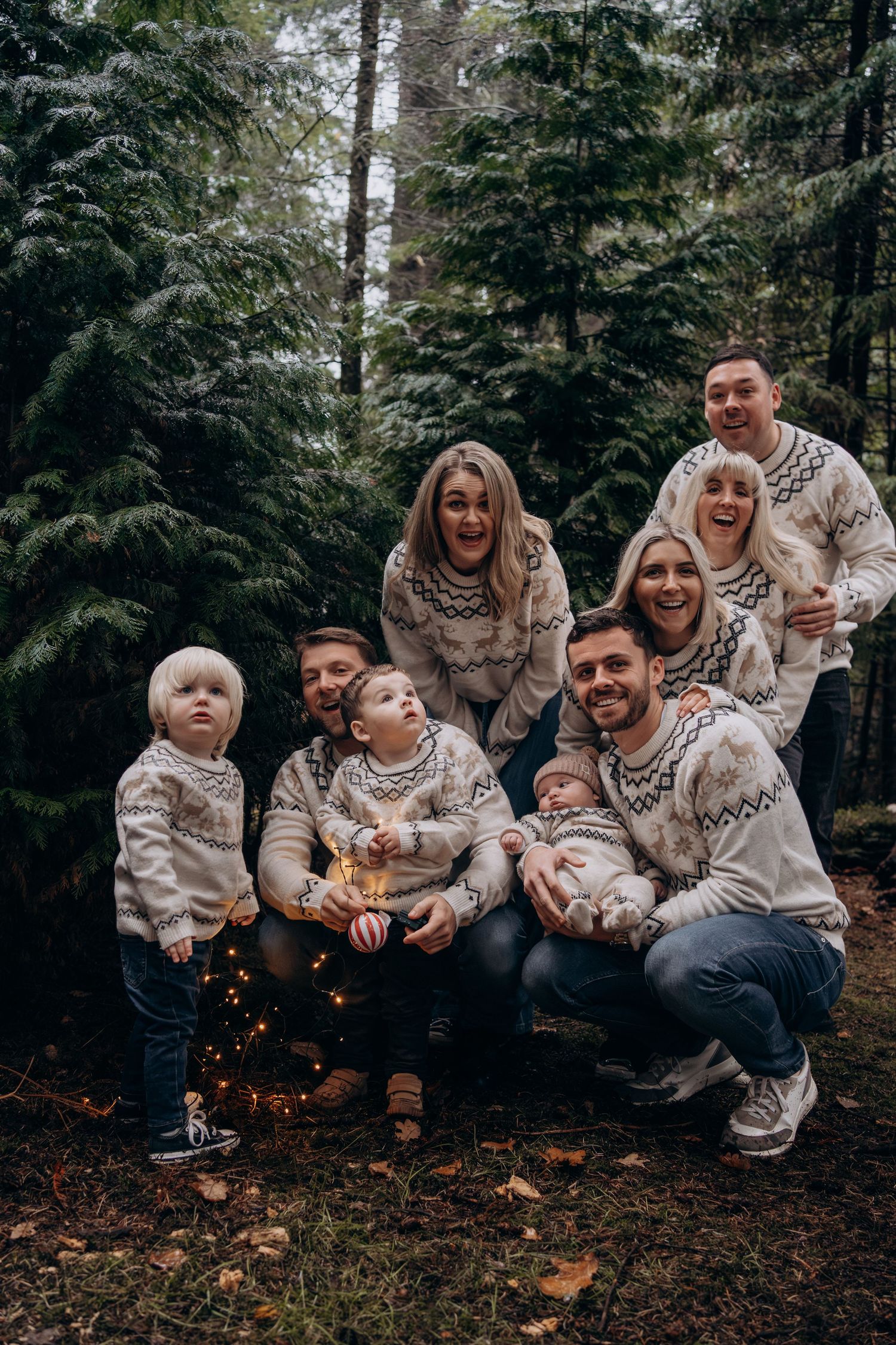 A family wearing matching cream sweaters poses together on fallen autumn leaves in a forest setting.