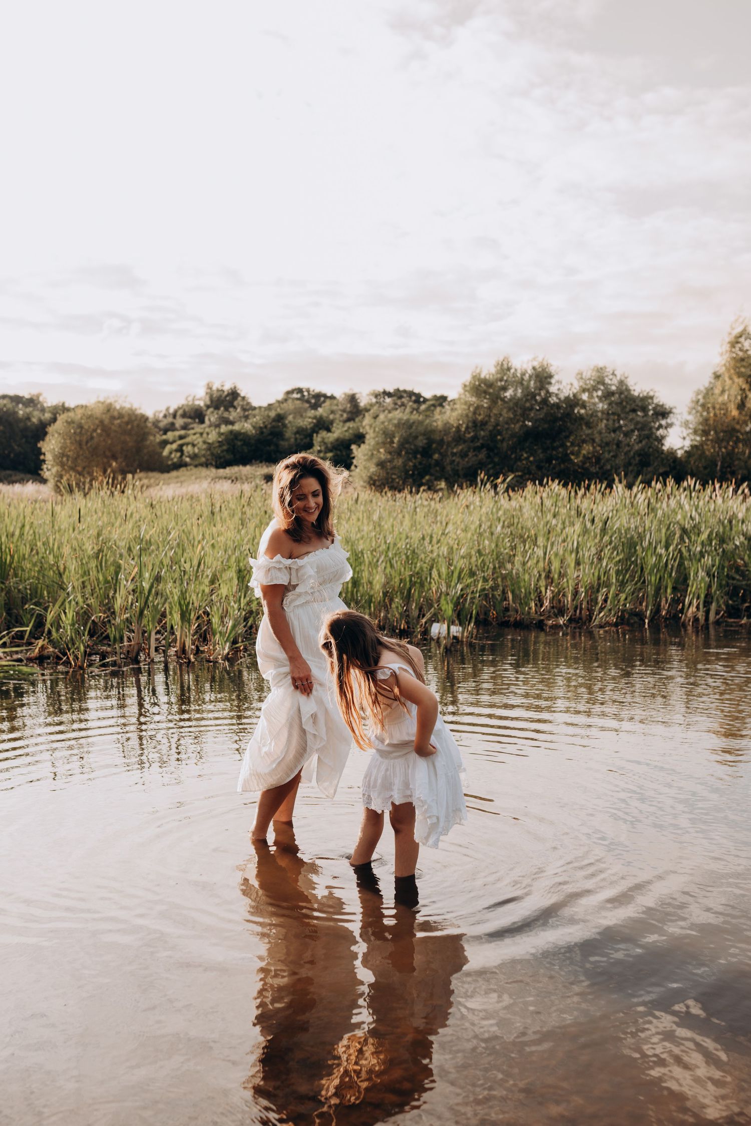 Two people in white dresses stand in shallow water creating ripples at sunset.