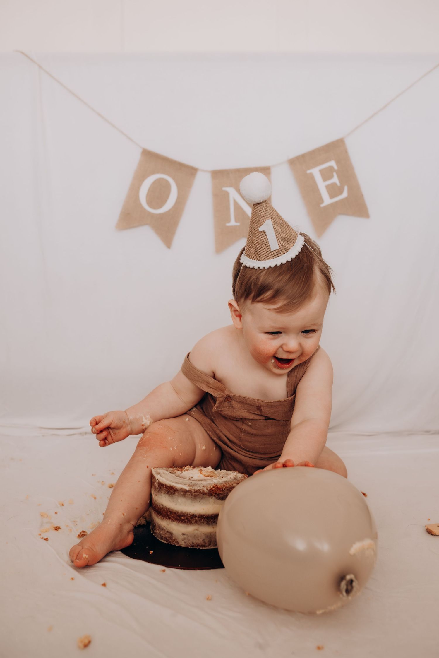 Baby in brown outfit smiling and playing with cake during a first birthday photoshoot.