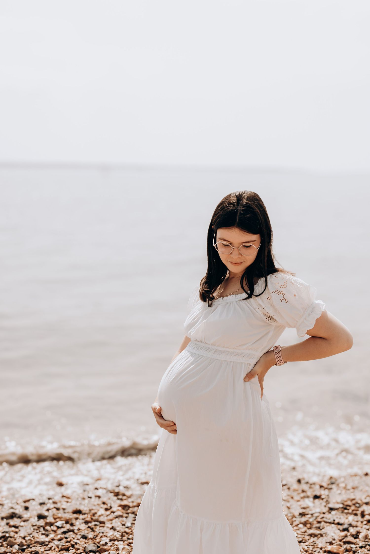 Expectant mother in white dress poses on beach during maternity photoshoot.