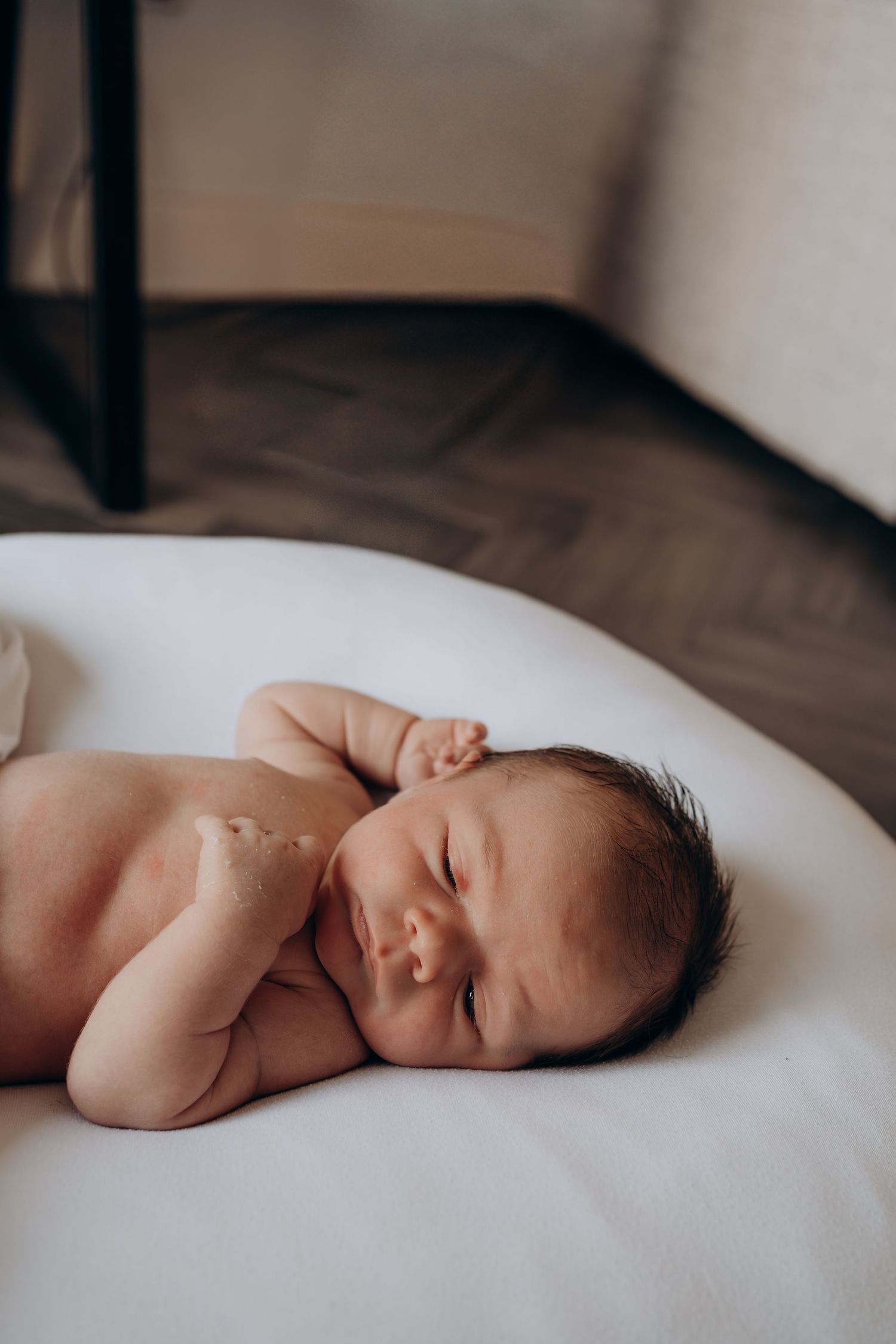 A peaceful newborn baby with dark hair sleeping curled up on a soft white surface.