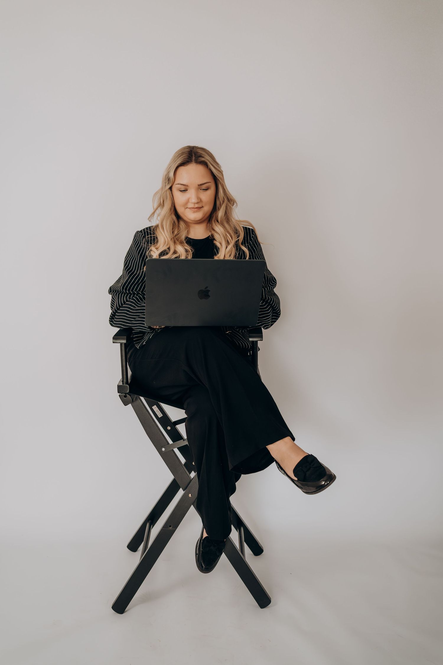 Woman in all black attire working on laptop while sitting on director's chair against light gray background.