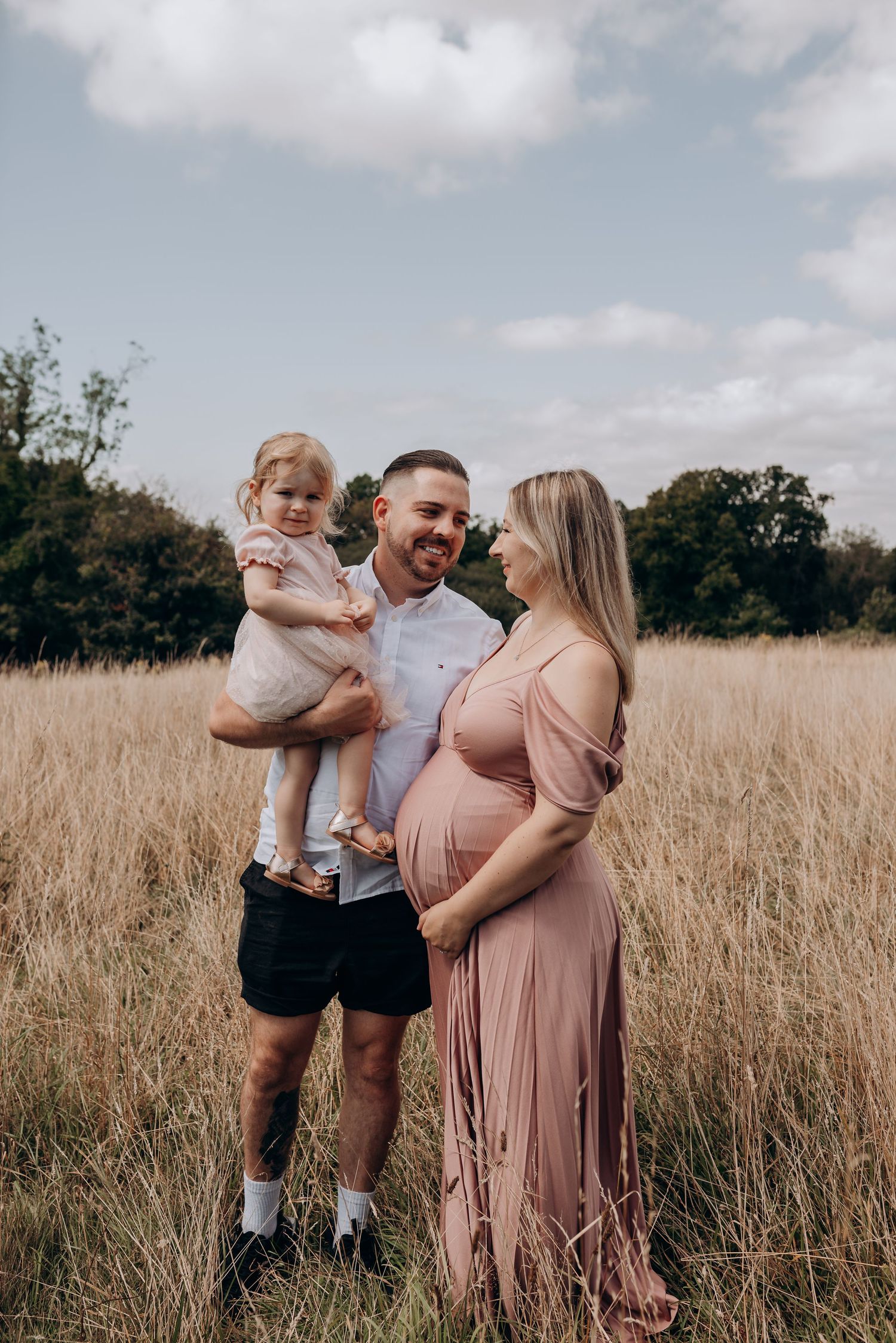 Family portrait in sunlit meadow with tall grass and cloudy sky background.