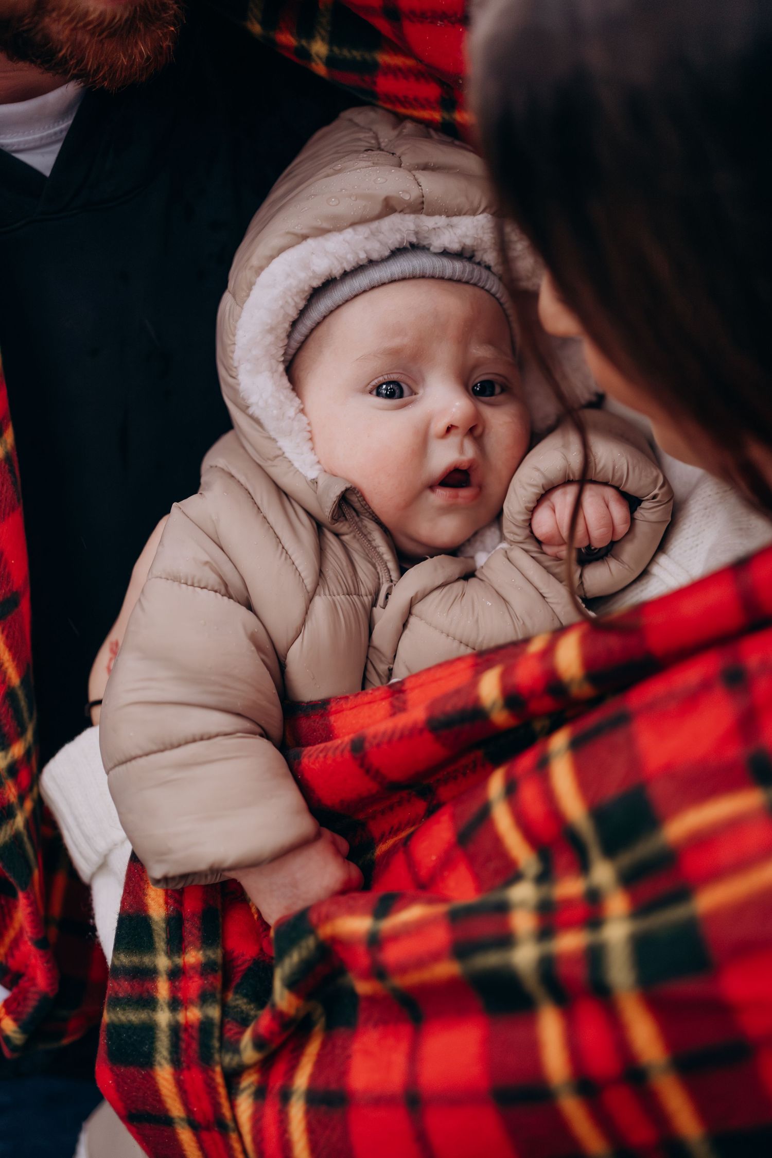 Baby in beige winter coat lies on red plaid blanket during cozy winter photoshoot.